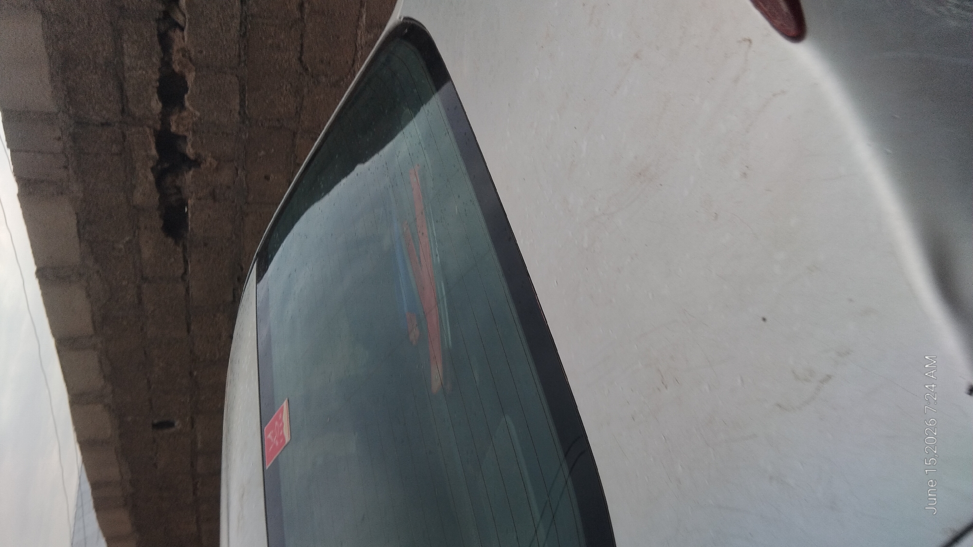 The image shows the rear windshield of a light-colored car parked in front of a brick wall. There is a red sticker on the upper left corner of the windshield and a red and blue object visible on the interior of the windshield. The car appears to be dusty. The brick wall is made of concrete blocks, and a dark, irregular opening is visible in the wall. The sky is overcast. The timestamp "June 15, 2026 7:24 AM" is visible in the bottom left corner. The location is identified as Howayi, Nigeria.