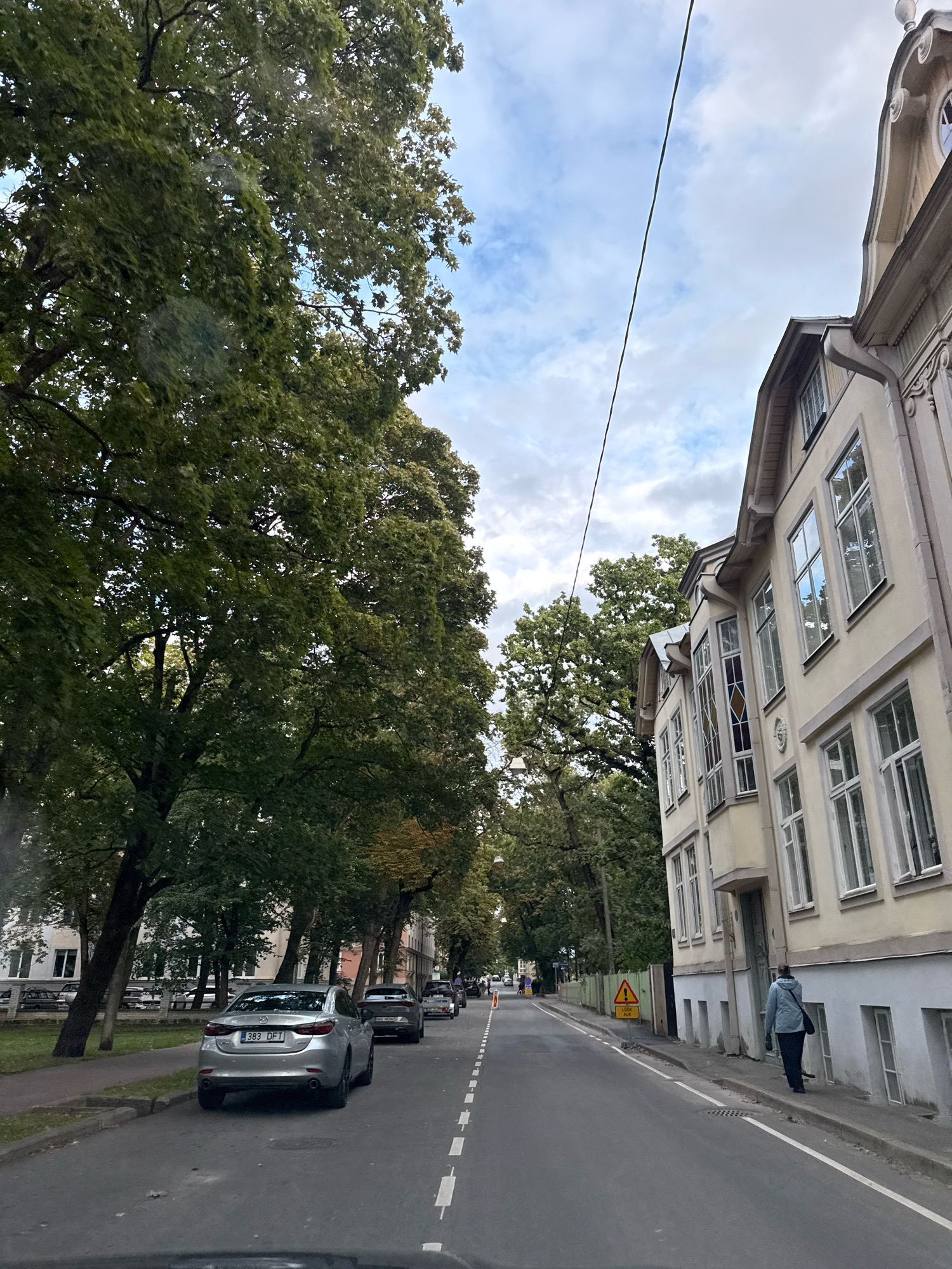 The image depicts a street scene in Tallinn, Estonia, likely during the daytime under an overcast sky. Tall trees with lush green foliage line the left side of the road, casting shadows. On the right, a light-colored building with multiple windows and ornate architectural details stands alongside the street. A single person in a light blue jacket is walking on the sidewalk in front of the building. Several cars are parked along the street, and a silver car with a license plate reading "383 DFT" is prominently featured in the foreground. The road itself has lane markings and is in good condition. A yellow traffic sign with a warning symbol is visible near the sidewalk. The overall impression is of a quiet, residential street within the city.
