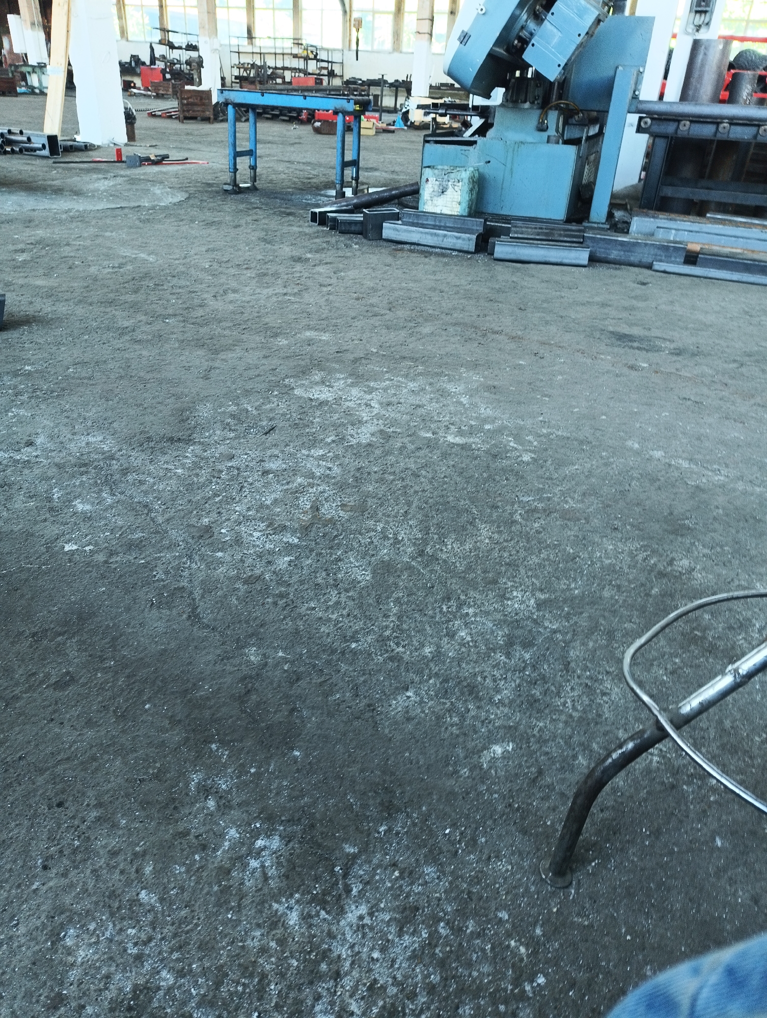 This image depicts an indoor industrial or workshop environment, possibly located in the city of Karnobat, Bulgaria. The ground surface is composed of coarse, uneven dirt or gravel, with scattered white debris. In the background, various metalworking machinery is visible, including a large blue cutting or bending machine. Metal pipes and components are also present, both on the floor and stacked. A blue metal stand or bench is situated in the mid-ground. The lighting suggests an interior space with daylight filtering through windows. In the foreground, a metal chair leg is visible, along with a portion of blue fabric, likely denim, indicating a person is present but not fully in view. The overall impression is of an active or recently used workspace.