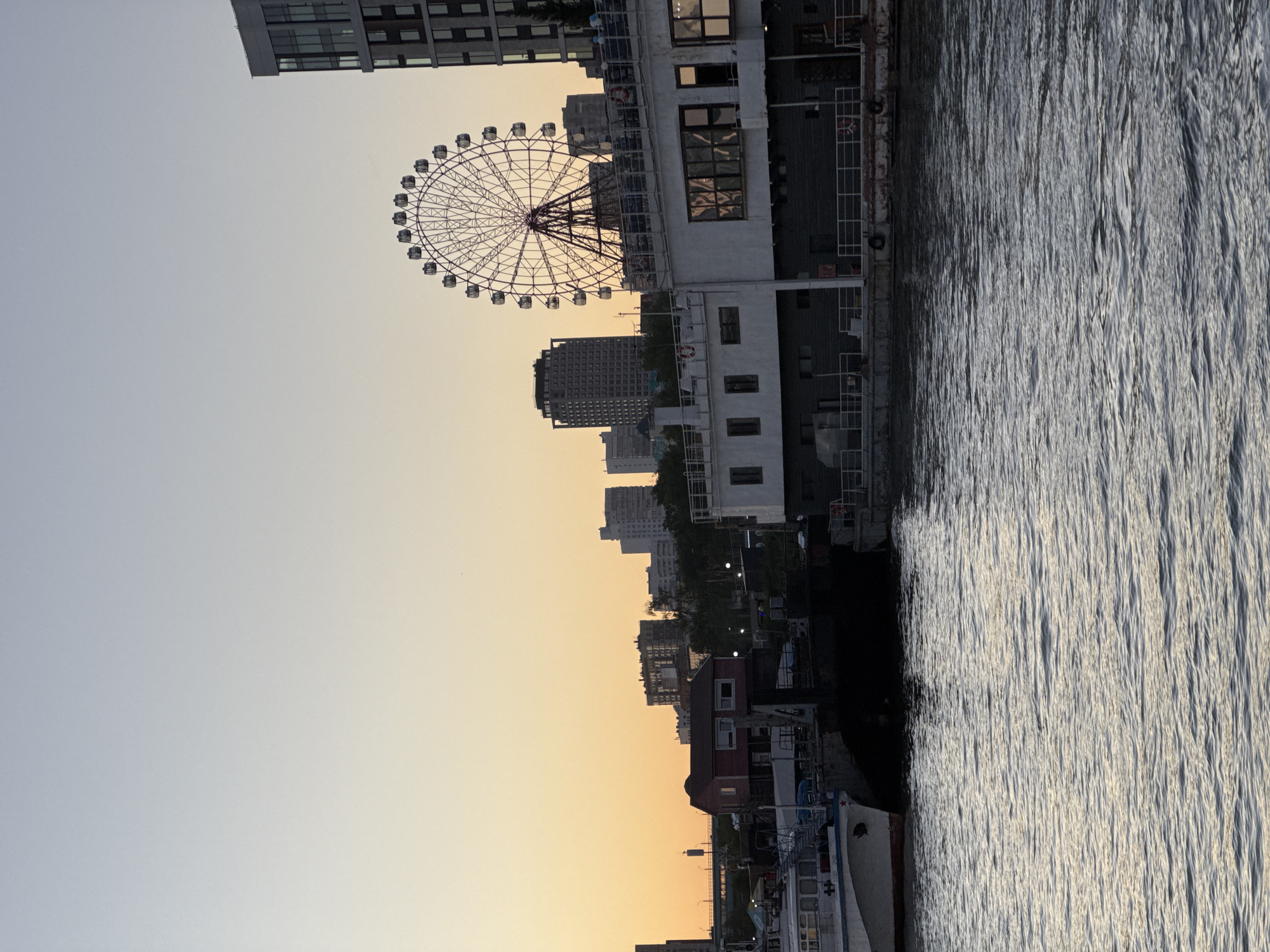 This is a rotated image of a waterfront cityscape at dusk in Novosibirsk, Russia. The foreground features choppy water with reflections of light. Along the shore, a variety of buildings are visible, ranging from modern, multi-story structures to smaller, older buildings and docked boats. A prominent Ferris wheel stands out against the skyline, suggesting a recreational area or amusement park. The sky transitions from a soft orange hue near the horizon, indicating sunset, to a pale blue overhead. The overall atmosphere is calm and serene, typical of an evening by the water. No people are clearly visible.