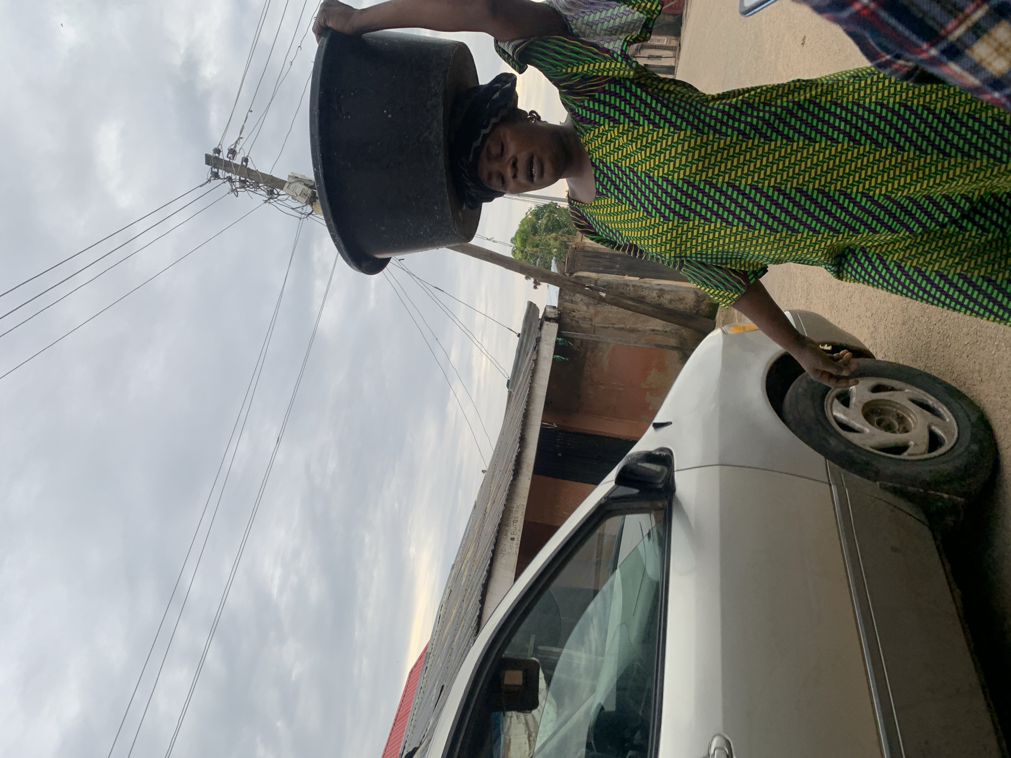 The image depicts an outdoor street scene in Jos, Nigeria, under an overcast sky. In the mid-ground, an adult female of African descent is seen walking from left to right. She carries a large, black, round basin on her head, supporting it with her raised left hand. She is dressed in a patterned garment featuring green and dark blue or purple zigzag designs and a dark headwrap. Her mouth is slightly open, suggesting exertion.
To the left of the woman, a light-colored (likely silver) minivan or large sedan is partially visible, showing its passenger-side front door, side mirror, and front wheel. In the background, multiple low-rise buildings with corrugated metal roofs are present. On the roof of one building, partially obscured by the vehicle, white text reads "YOU CAN BUY SHEA BUTTER HERE." Overhead, a wooden utility pole with a complex network of electrical wires extends into the cloudy sky. Several parallel power lines stretch across the upper portion of the frame. The ground appears to be unpaved. A hand with a plaid shirt sleeve is partially visible on the far right edge of the frame.