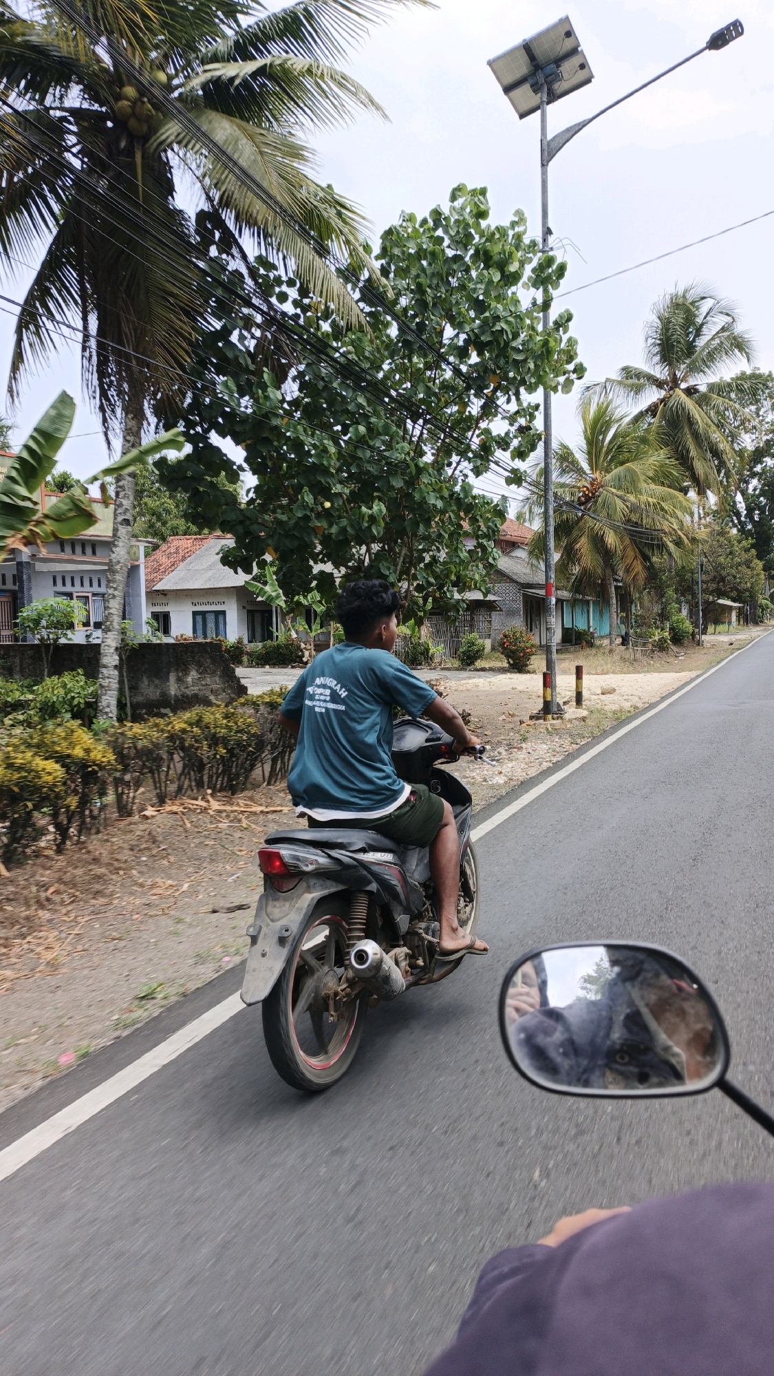 In the city of Cibenda, Indonesia, a person is riding a motorcycle on a paved road. The rider, seen from behind, is wearing a teal t-shirt and dark shorts. They are operating a black motorcycle with a red rim on its wheel. In the foreground, the handlebar and rearview mirror of another motorcycle are visible, reflecting the sky and trees. The surrounding environment features houses with red-tiled roofs, lush green trees, and palm trees, indicating a tropical or subtropical climate. A solar-powered street light stands tall on the side of the road. The road itself has a white line marking its edge. The time of day appears to be daytime, with overcast or partly cloudy skies.