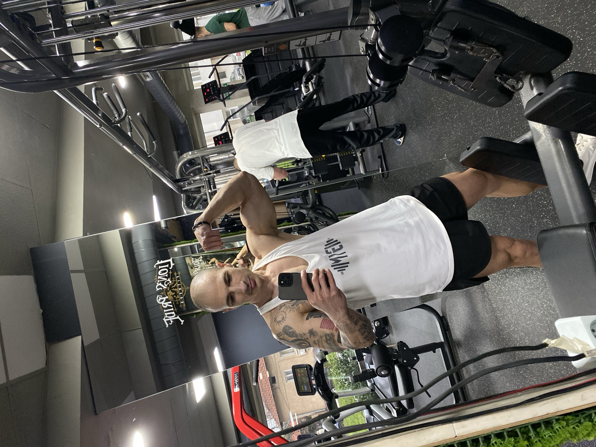 The scene depicts the interior of "Lion's Pride Gym" in Pleven, Bulgaria, captured via a mirror reflection. A male individual, appearing in his 30s or 40s, with a shaved head and tattoos covering his left arm, is positioned slightly off-center to the left. He wears a white sleeveless tank top with a stylized barbell logo on the chest and black athletic shorts. He is flexing his left bicep while holding a dark-colored smartphone in his right hand, angled towards the mirror, indicating he is taking a selfie. His gaze is directed towards the phone screen. White athletic shoes are visible on his feet.
In the background, visible in the mirror, are other gym patrons and equipment. To the right of the primary subject, another male, dressed in a white t-shirt and black athletic pants, is seen from behind, positioned near a cable machine. Further right, a third male in a green t-shirt and light gray pants is partially visible. The gym floor is covered with black speckled rubber matting. Various exercise machines are present, including multiple black-framed cable machines with weight stacks, and two treadmills, one prominently red-framed, each with digital displays. A leg curl/extension machine with black padding is in the immediate foreground. The ceiling is dark grey with visible ventilation ducts and recessed lighting. A white electrical outlet with black and red taped wires is visible in the lower left, adjacent to a textured green wall panel. Through a large window, residential buildings with tiled roofs and outdoor vegetation are visible, indicating an urban setting.
