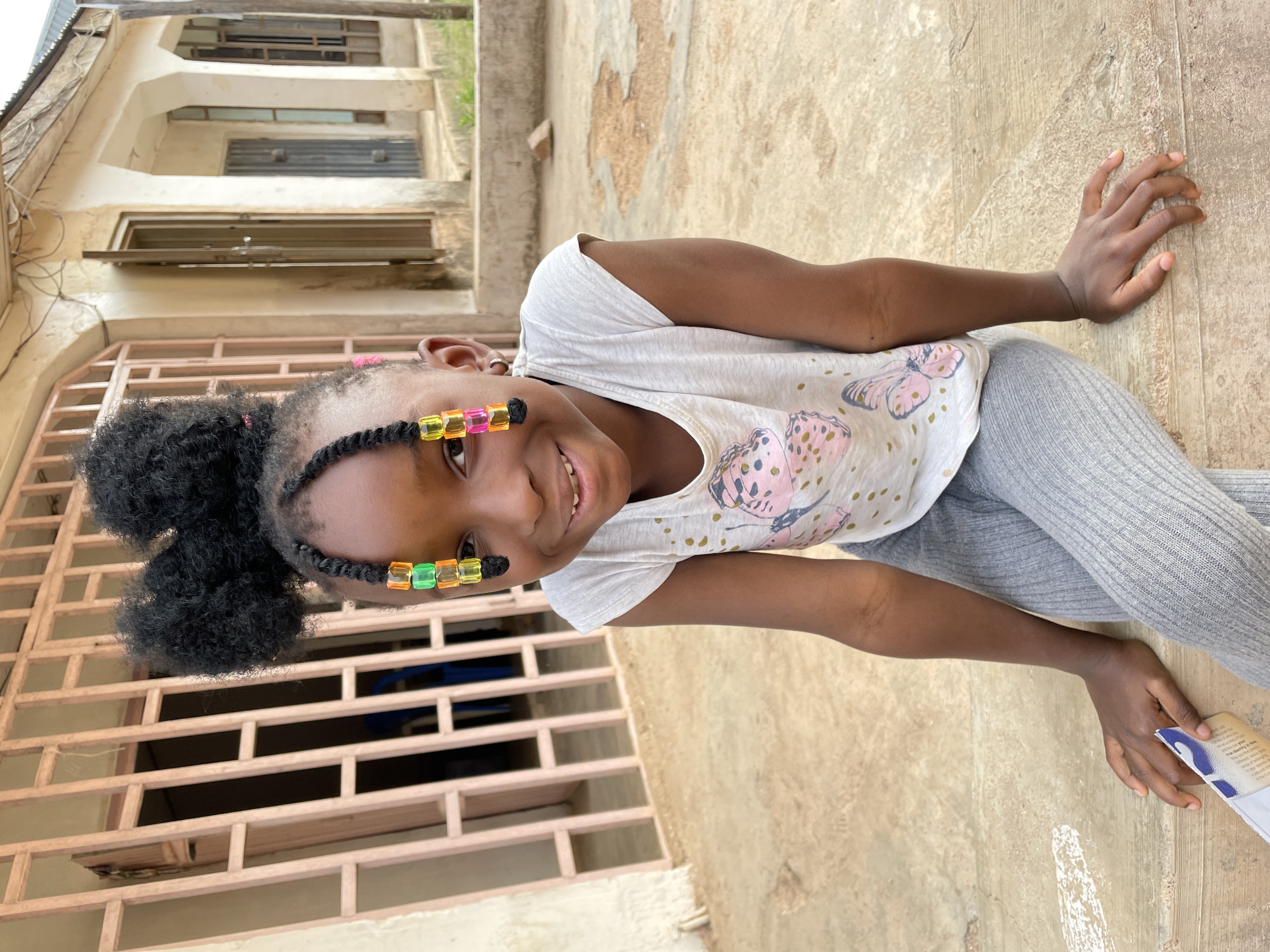 A young Black girl is captured smiling warmly at the camera in a sideways-oriented outdoor full-shot. She is sitting or kneeling on a light-colored, paved surface, with her left hand resting on the ground. Her dark, curly hair is styled in two small pigtails, and her front braids are adorned with colorful square beads in shades of yellow, orange, and green.
She is wearing a light-colored short-sleeved t-shirt featuring multiple pink and patterned butterfly designs, paired with grey ribbed leggings. Her right hand holds a small, light-colored object, possibly a card or booklet, on which some English text is partially visible, including phrases like "...what do you...", "...that is what you...", "...look closely to him...", "...products and services...", and "...d by the Creator".
The background features a multi-story building with light-colored, possibly beige or off-white, walls showing some signs of wear. Several windows with dark shutters or open frames, and metal security grilles or railings, are visible. The ground is a solid, light-colored material, likely concrete or tile. The scene is brightly lit, indicating daytime, and the girl's expression conveys happiness and engagement. The architecture and outdoor setting suggest a residential area, consistent with the specified location of Barakin Akawo, Nigeria.