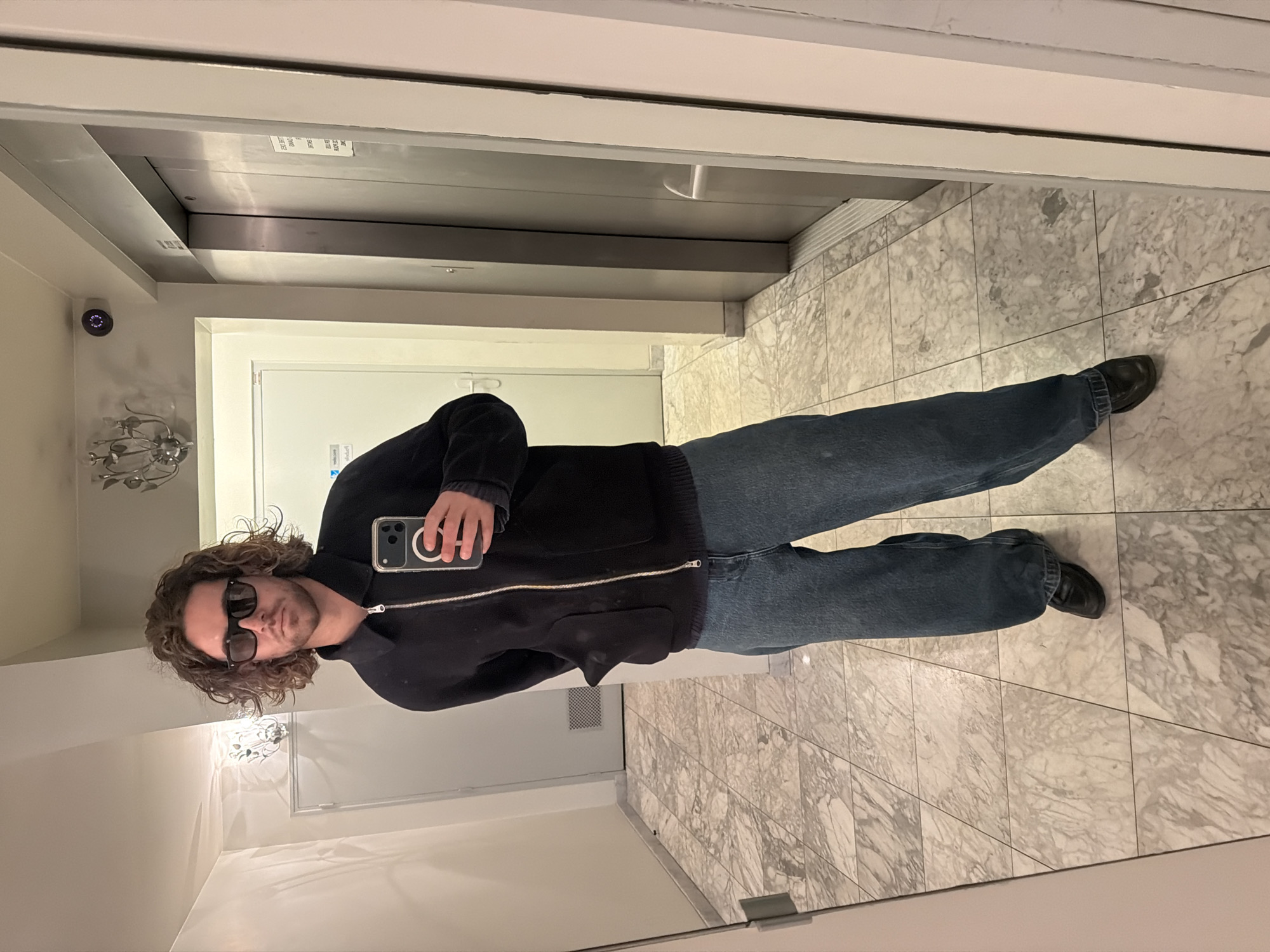 A person with curly brown hair and sunglasses is taking a selfie in a mirrored space. The person is wearing a dark-colored jacket and blue jeans, and black shoes. The background includes a tiled floor, white walls, light fixtures, a door, and an elevator. There is no verifiable information indicating the location is Paris, France.
