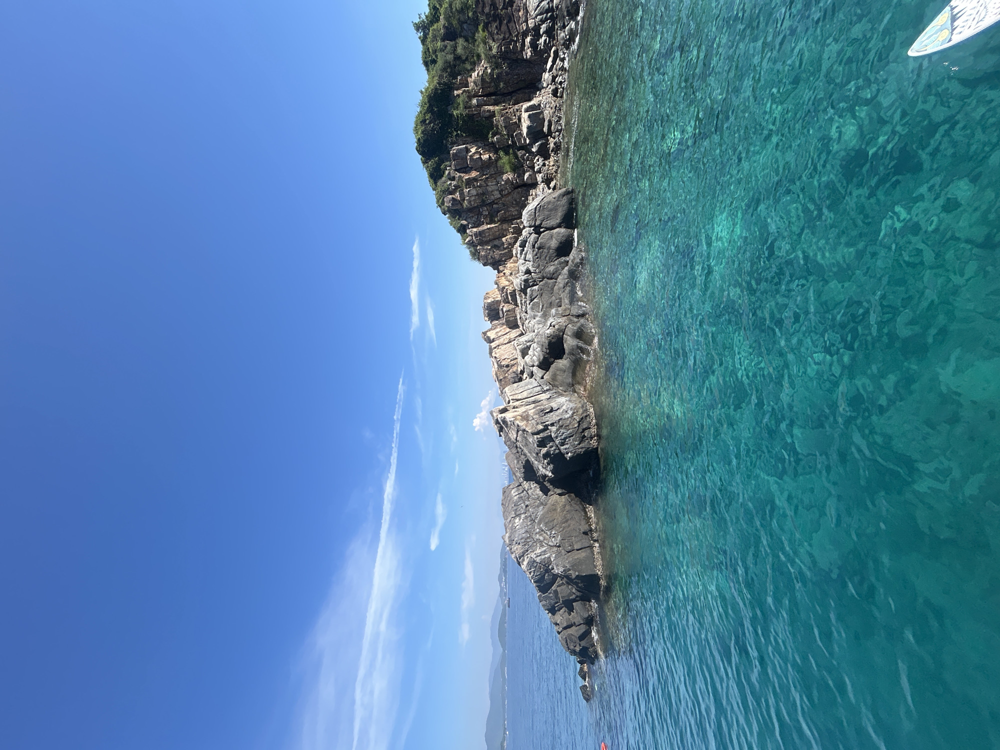 The image shows a scenic view of a rocky coastline with clear, turquoise water in the foreground. The water is so transparent that the seabed is visible in places. The rocks are jagged and appear to be part of a small island or peninsula. Lush green vegetation covers the top of the rocky landmass. In the distance, hazy mountains can be seen along the coastline, and the sky is a clear blue with a few wispy white clouds. The scene evokes a sense of natural beauty and tranquility, characteristic of tropical or subtropical coastal regions. This particular location is identified as being in the city of Bac Nha Trang Ward, Vietnam, a coastal area known for its beaches and islands.