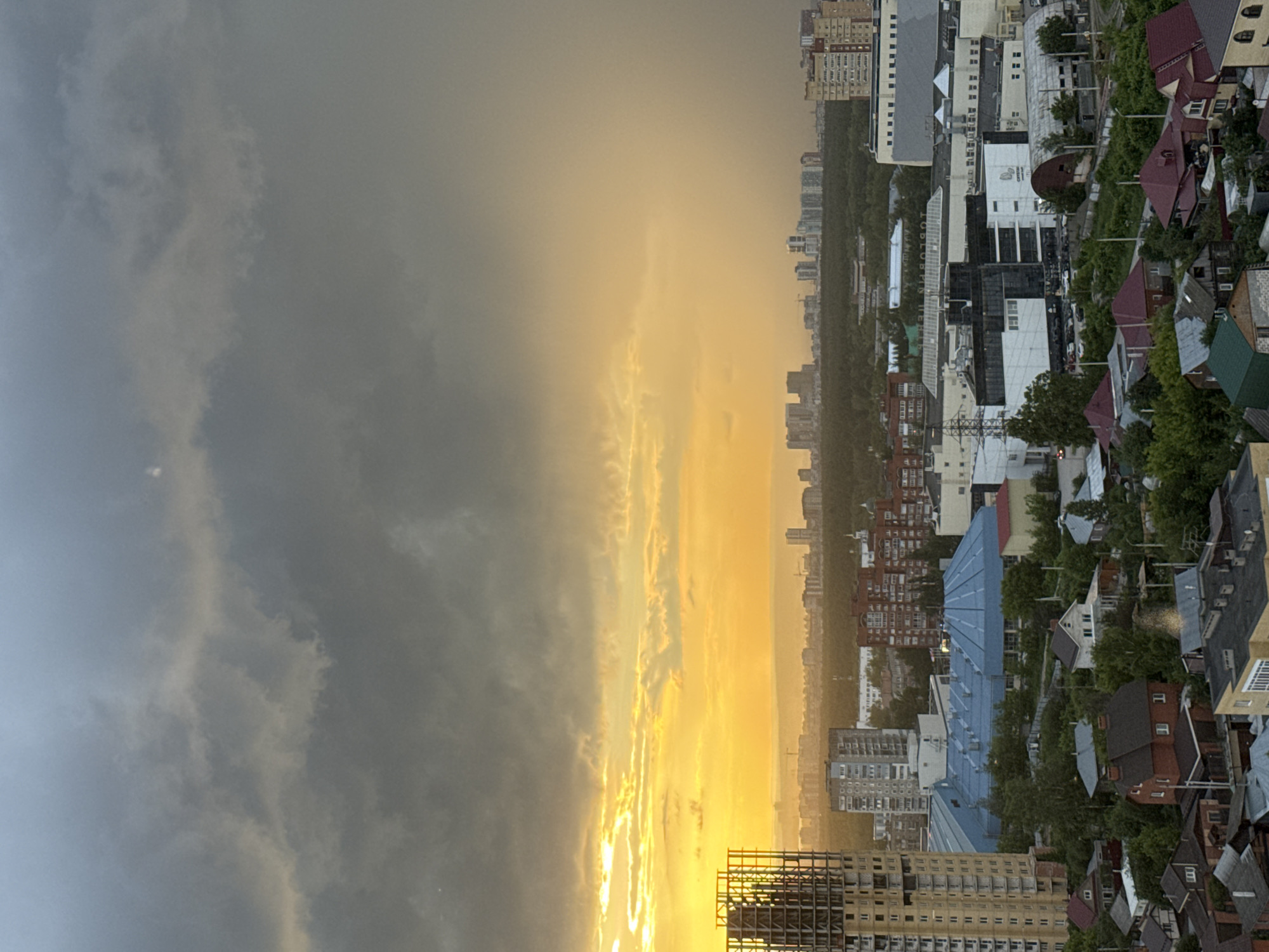 The image depicts an aerial view of a city under a dramatic sky. The sky is filled with dark, heavy clouds, particularly on the left side, with a bright, golden-yellow light breaking through from the horizon on the left. This suggests either a sunrise or sunset, with the sun's rays illuminating the clouds and casting a warm glow over a portion of the cityscape.
In the foreground, a skeletal structure of a tall building under construction is visible on the left. Below and to the right of this, the city unfolds with a mix of buildings. There are several multi-story residential buildings, some with red brick facades and others with lighter colored exteriors. A large building with a distinctive blue, domed roof is also prominent. Further into the city, more urban development can be seen, with a cluster of taller buildings in the distance, some appearing to be modern high-rises.
Interspersed with the urban structures are areas of dense green trees and vegetation, suggesting parks or forested areas within or bordering the city. In the lower right of the image, a denser residential area with smaller houses and rooftops is visible.
The overall impression is one of an active urban environment at the cusp of changing weather conditions, with the dramatic sky creating a strong visual contrast with the organized structures of the city. There are no visible people or actions occurring in the image. The location is identified as Shirkovo, Russia.