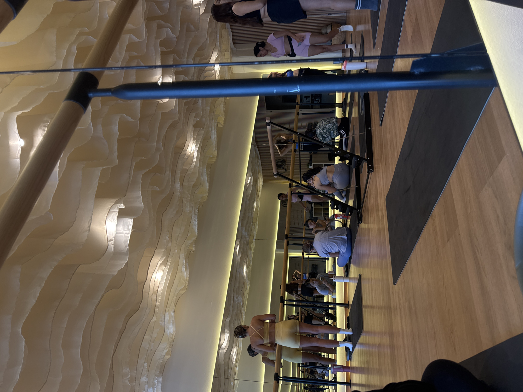 A group of people are engaged in a fitness activity, possibly Pilates, in what appears to be a studio setting. Several individuals are positioned along a ballet barre, with some performing exercises on mats. The environment includes a large mirror along one wall, reflecting the participants and amplifying the sense of space. The studio features unique wall decorations, with layered, semi-transparent materials illuminated by internal lighting. The location is likely an upscale fitness studio in San Bernardino Tlaxcalancingo, Mexico, based on the architectural style and the presence of the barre and mats.