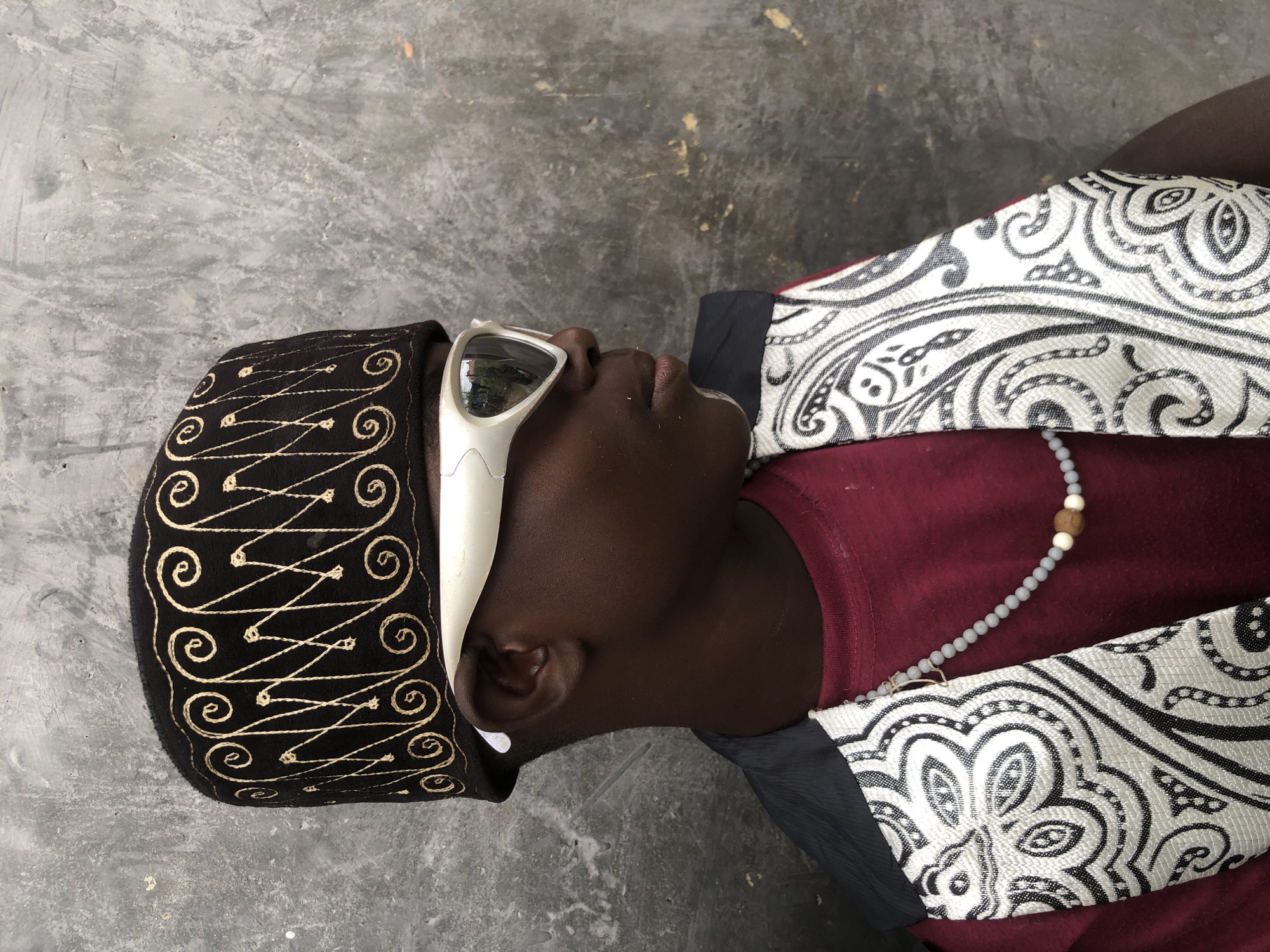 The image is a portrait of a young Black person wearing a dark, embroidered kufi hat, white futuristic-style sunglasses, a maroon t-shirt, and a patterned vest. The person is positioned in profile, facing to the right, against a rough, gray concrete wall. The wall shows variations in texture and color, with some lighter patches and darker smudges. The person's dark skin contrasts with the lighter tones of the sunglasses and the patterned vest, which features intricate black and white paisley-like designs. A beaded necklace with a few wooden beads is visible around the person's neck. The lighting appears to be natural, coming from the front and side, casting subtle shadows. The background suggests an outdoor urban environment, potentially in a courtyard or alley. This scene is set in the city of Ado, Nigeria.