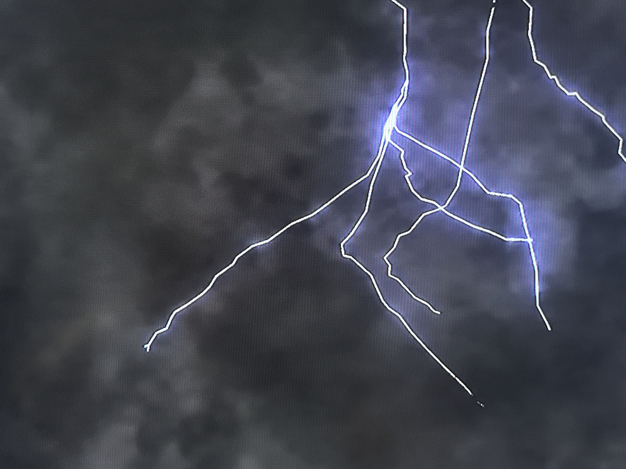 The image presents a dramatic view of a powerful lightning strike against a dark, stormy sky, likely in Varna, Bulgaria. The central subject is a brilliant, branching lightning bolt, primarily depicted in electric blue and bright white. Its core emanates an intense glow where multiple branches converge, and numerous thinner, intricate tendrils extend across the frame, illuminating the surrounding clouds.
The sky is filled with heavy, dark gray clouds, indicating a severe thunderstorm. The low light conditions and the vivid illumination from the lightning suggest that the event is occurring at night or during a heavily overcast day. The overall atmosphere conveyed is one of raw natural power and intensity.
There are no people or discernible man-made objects visible in the image, keeping the focus entirely on the meteorological phenomenon. No visible text is present. A subtle, fine grid-like pattern is noticeable across the entire image, suggesting that it might be a photograph of a digital screen or display. The scene captures the awe-inspiring and formidable beauty of a lightning storm.