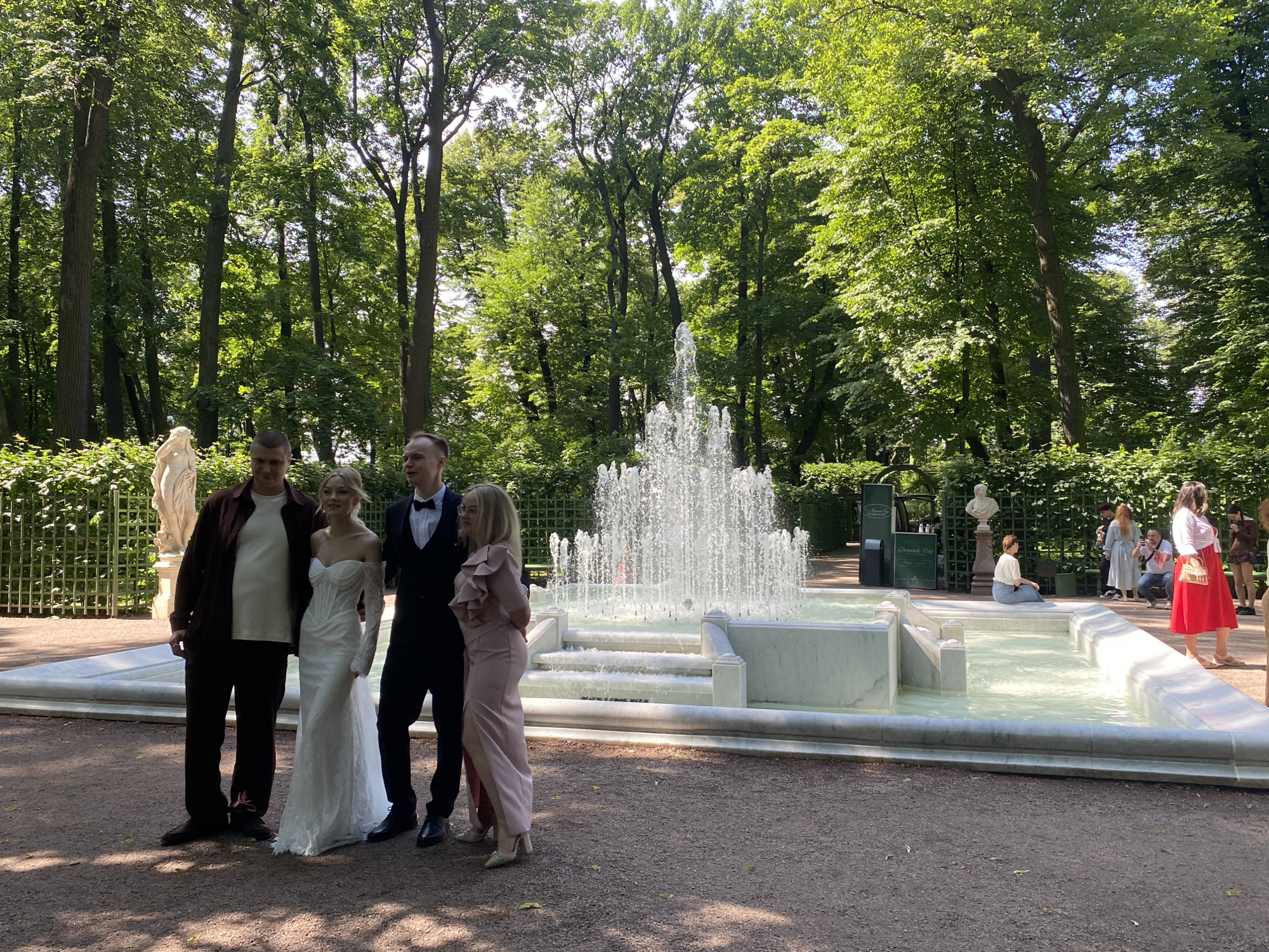 In Saint Petersburg, Russia, four individuals stand posed in front of a multi-tiered fountain in a sun-dappled park setting. A man in a dark brown jacket and white t-shirt stands to the left, next to a woman in a white wedding dress with long sleeves and a sweetheart neckline. To their right is a man in a black suit with a bow tie, and beside him, a woman in a pale pink off-the-shoulder dress with ruffled details and a high slit. The fountain, constructed of white marble, features water spraying upwards in a central plume and cascading down the tiers. The background is dominated by lush green trees, with glimpses of statues and ornate fencing. Other individuals can be seen in the background, some observing the fountain, and one person is seated by the water's edge. The ground is covered in gravel.