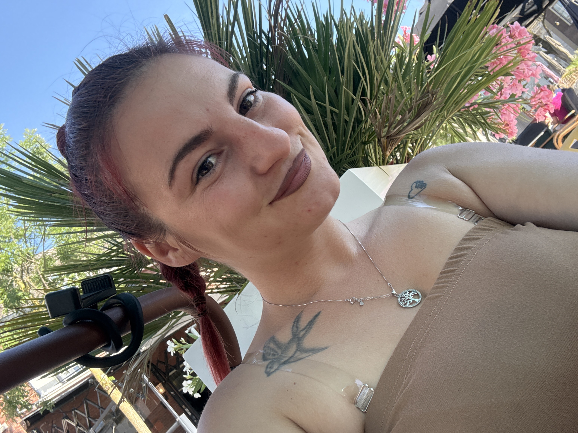 In an outdoor setting, possibly a patio or balcony, a young woman with reddish-brown hair tied in a braid is taking a selfie. She is wearing a tan strapless top and a silver necklace with a tree of life pendant. Tattoos are visible on her shoulder and upper chest. Behind her, palm fronds and pink flowers suggest a warm climate, and a structure resembling a building facade can be seen in the background. The lighting indicates it is daytime, likely sunny. The scene does not contain any elements that place it specifically in Veghel, Netherlands.