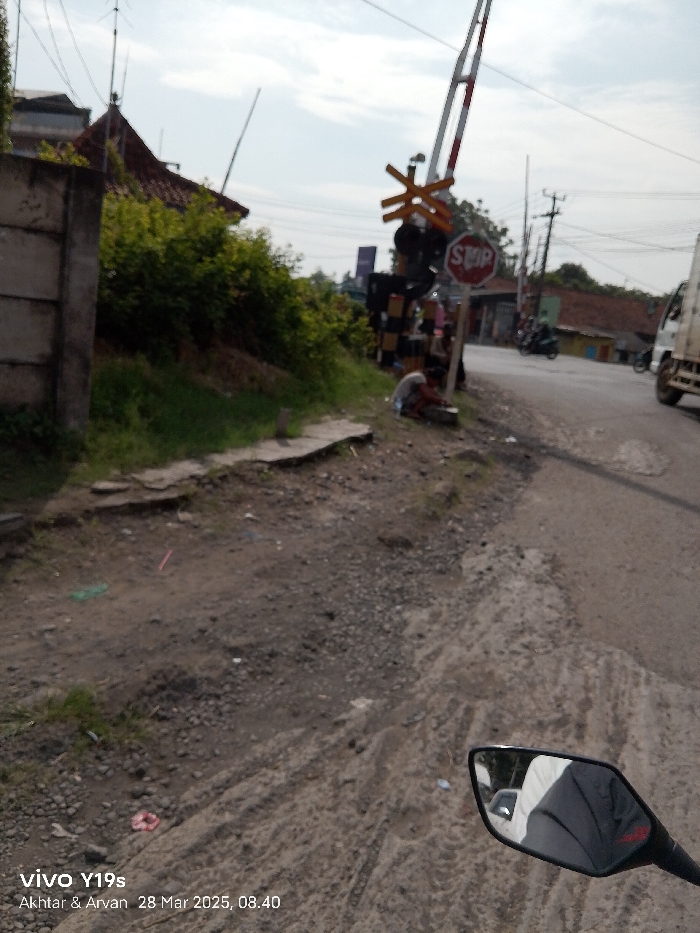 The image shows a view from a motorbike, facing a railroad crossing in Jatibarang, Indonesia. The crossing gate is down and a red "Stop" sign is visible. A person is sitting on the ground to the left of the sign. The road is paved with asphalt and there is a dirt shoulder on the left. The background shows a residential area with a few houses and trees. The motorcycle is moving slowly toward the crossing. The time is 08:40. The image appears to be taken by a passenger on a motorbike.