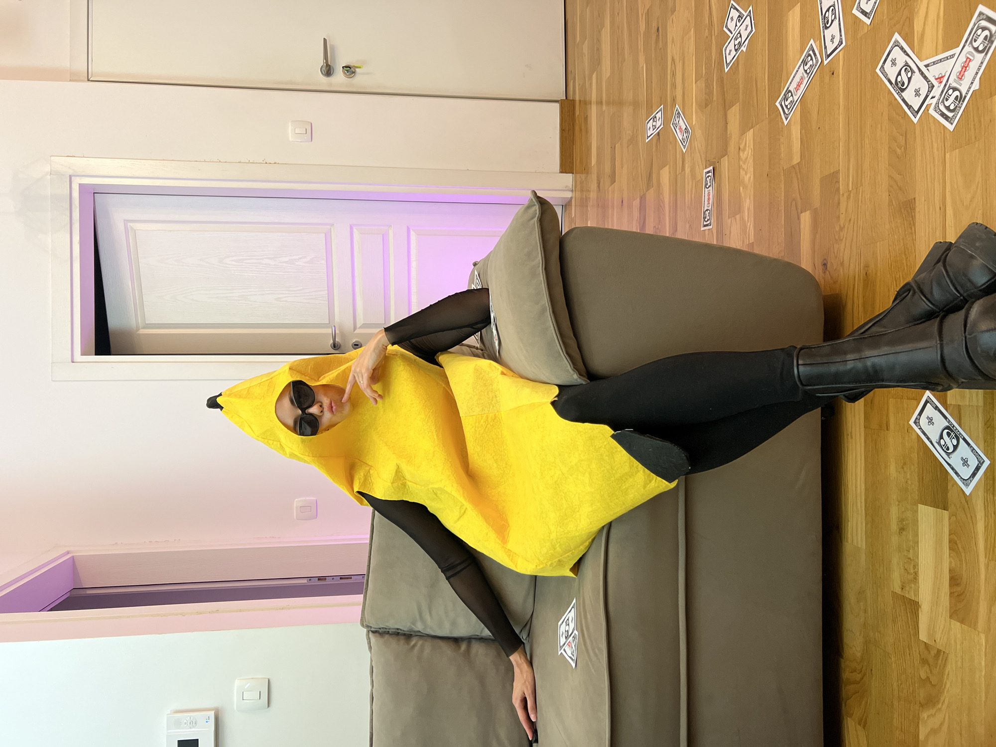 A person is depicted lying on a grey upholstered sofa in an indoor setting. The image is oriented 90 degrees counter-clockwise, giving the impression that the person is vertically positioned on the couch. The individual is wearing a vibrant yellow banana costume over a black long-sleeved top, paired with black leggings and black boots. They are also wearing black sunglasses and are posing for the camera with a relaxed and confident expression.
The sofa rests on a light wooden parquet floor. Numerous paper cutouts, designed to resemble play money, are scattered across the floor and partially on the sofa. Each cutout features a prominent dollar sign ($) and the visible text "S SUPER-PLAY".
In the background, a white wall with a decorative doorframe is visible. A white paneled door is slightly ajar, revealing a dark interior space from which a soft purple-pink light emanates. Above the doorframe, there's another section of white wall, an electrical switch or thermostat, and metallic door handles or hooks on what appears to be an overhead cabinet. The scene is brightly lit, with the colored light adding an atmospheric and playful element.