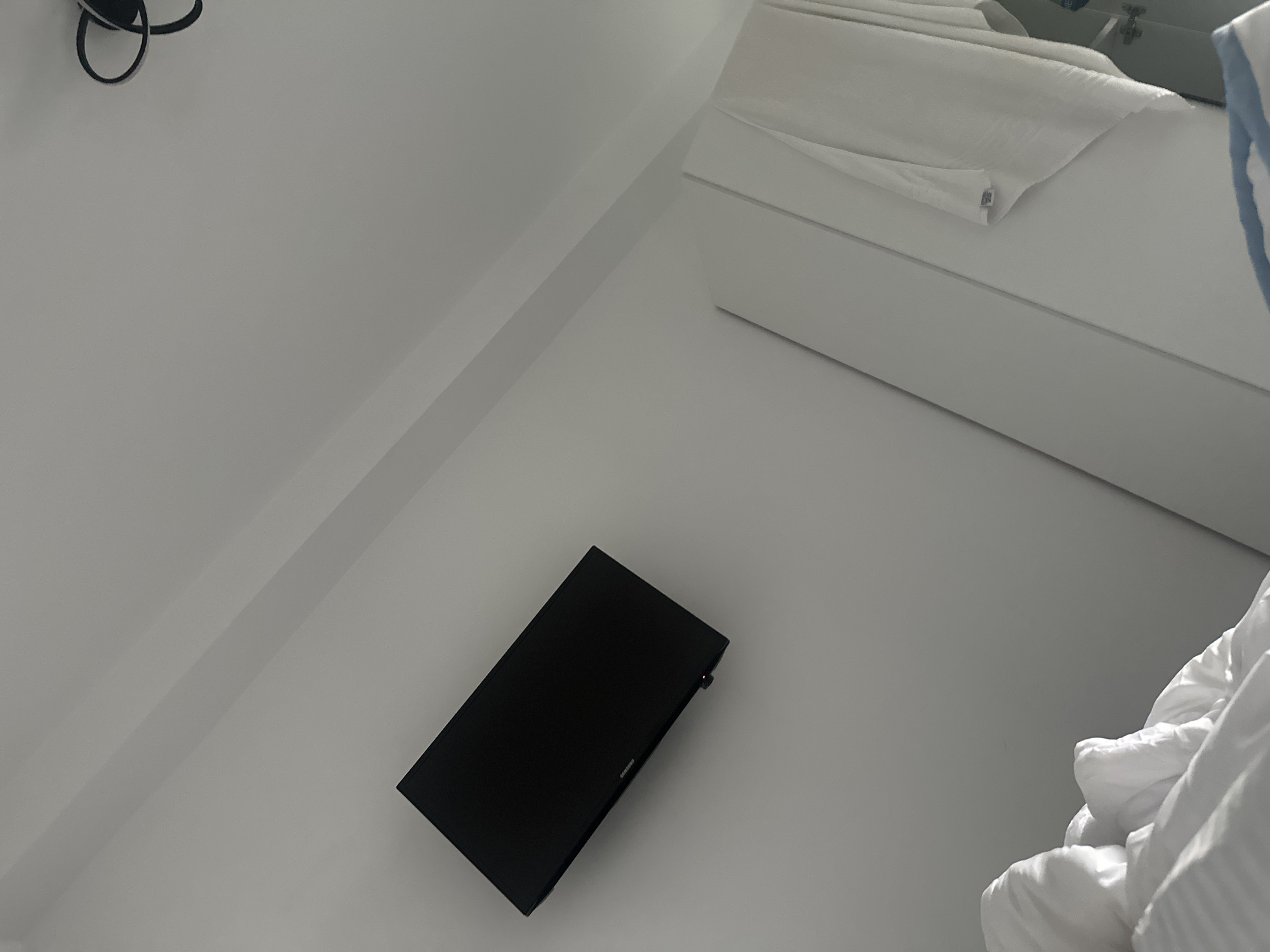 The image depicts an interior scene, likely a bedroom, angled to emphasize a wall-mounted black television. The television is positioned on a white wall, with a dark border and a small red indicator light visible at the bottom. To the right of the television, a portion of a white closet or wardrobe is visible, with a folded white towel or garment hanging from it. In the lower left corner, white bedding, possibly a duvet and pillows, is partially visible. The ceiling above is white and features angled sections. A black decorative light fixture is visible in the upper right corner. The scene appears to be clean and well-lit, with natural light casting shadows. The location is identified as Stațiunea Zoologică Marină Agigea, Romania.