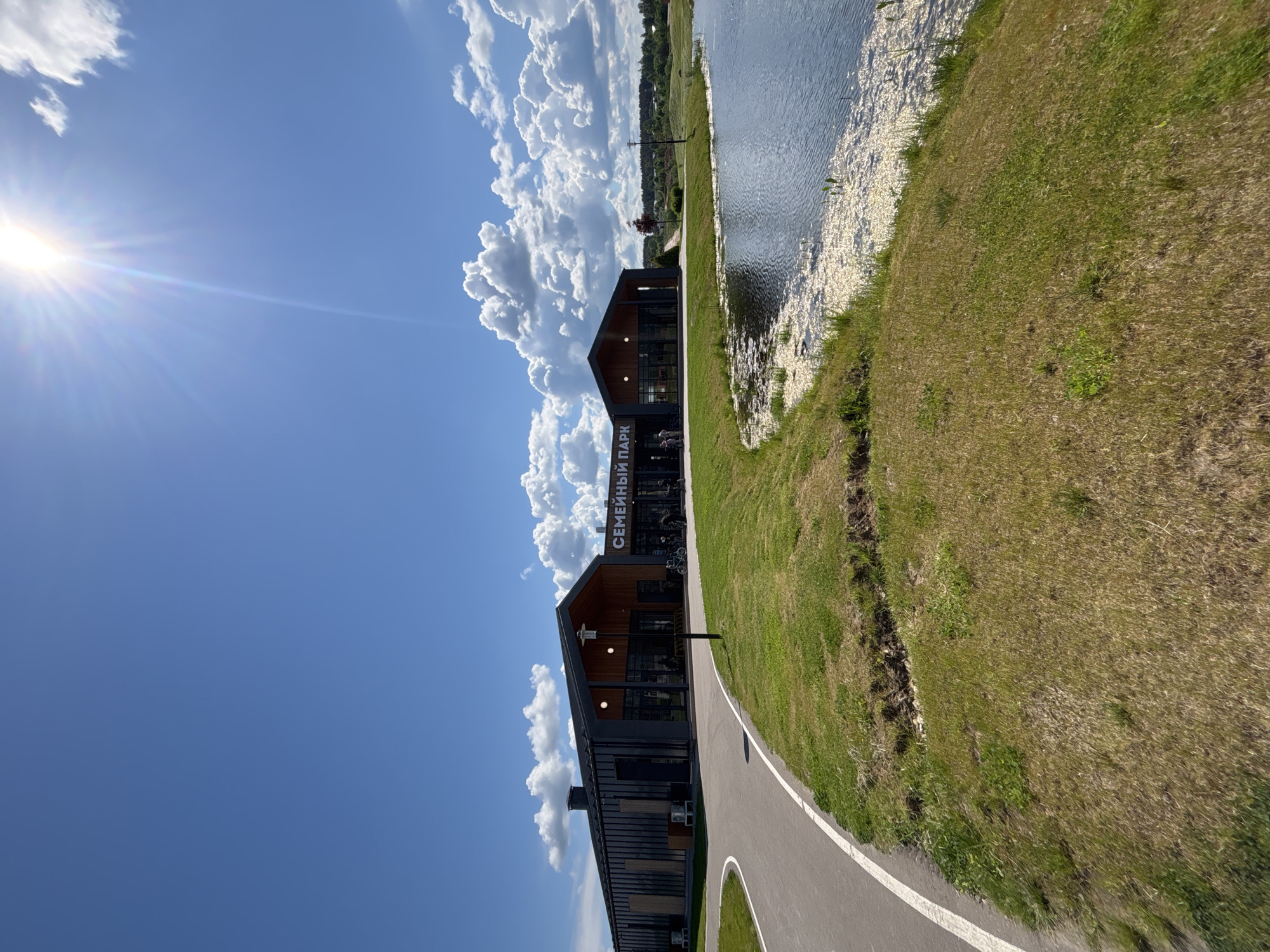 The image presents a scene at the “Семейный Парк” (Family Park) in Varaksino, Russia. The sky is partly cloudy, with bright sunshine. A paved path runs alongside a grassy area, with a body of water visible in the background, potentially a lake or river. The park building, with a dark exterior and a visible sign reading “Семейный Парк”, sits next to the path. The overall environment suggests a recreational area by the water on a sunny day.