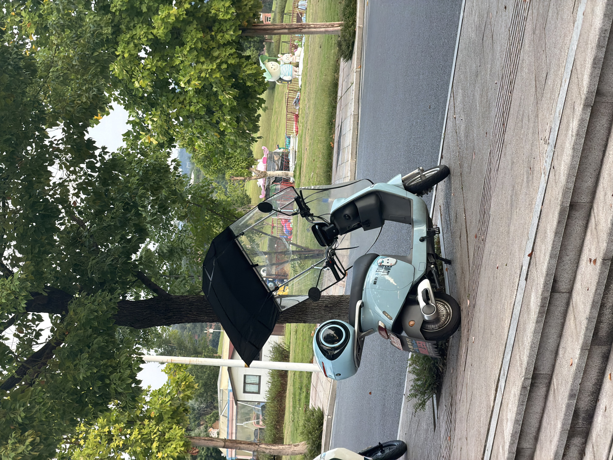 In the city of Jiangjin, China, a light blue electric scooter with a black awning and a clear windshield is parked on a sidewalk. The scooter is equipped with a spherical, light blue storage box on the rear. A small bush is located behind the rear wheel of the scooter. The sidewalk is adjacent to a road and a grassy park area. In the park, a statue of a cartoon bear in a striped shirt and a hat, accompanied by a rabbit, stands on the grass. Further in the background, there are trees, a building with a red roof, and what appears to be playground equipment. The scene is captured during the day, with trees and foliage prominently featured in the foreground and mid-ground.