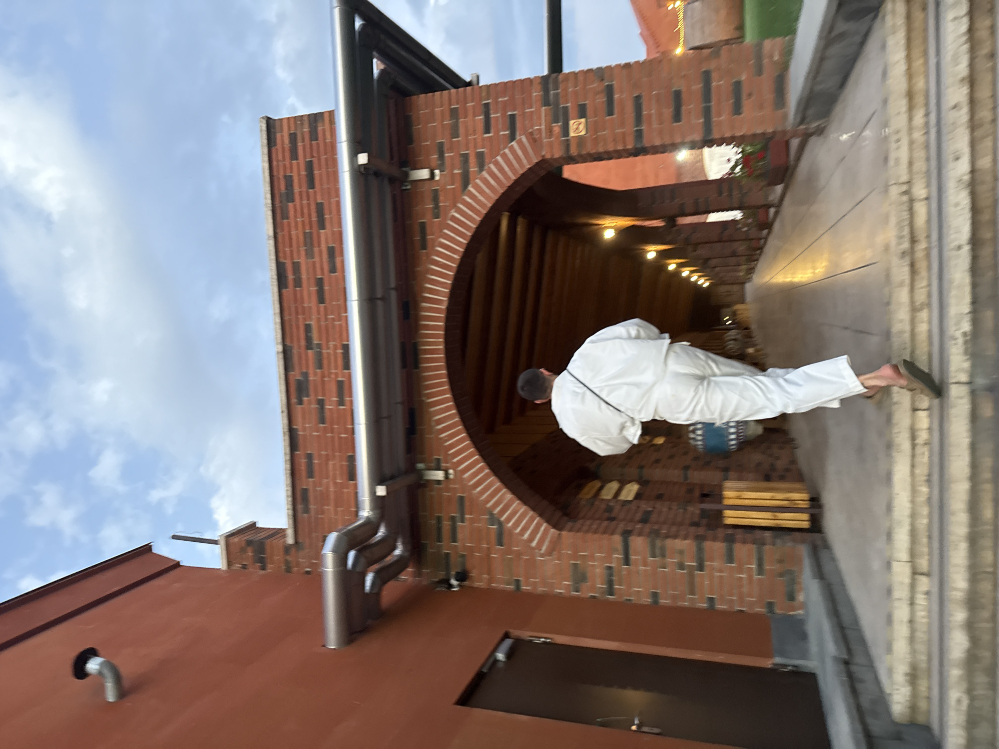 The image depicts a person wearing white clothing walking up a set of stairs towards a brick archway. The archway leads into a covered walkway illuminated by lights. The building is constructed of red brick and has a large brown door on the left. A drainage pipe runs along the side of the building. The sky is cloudy. The location is identified as Sasunik, Armenia.