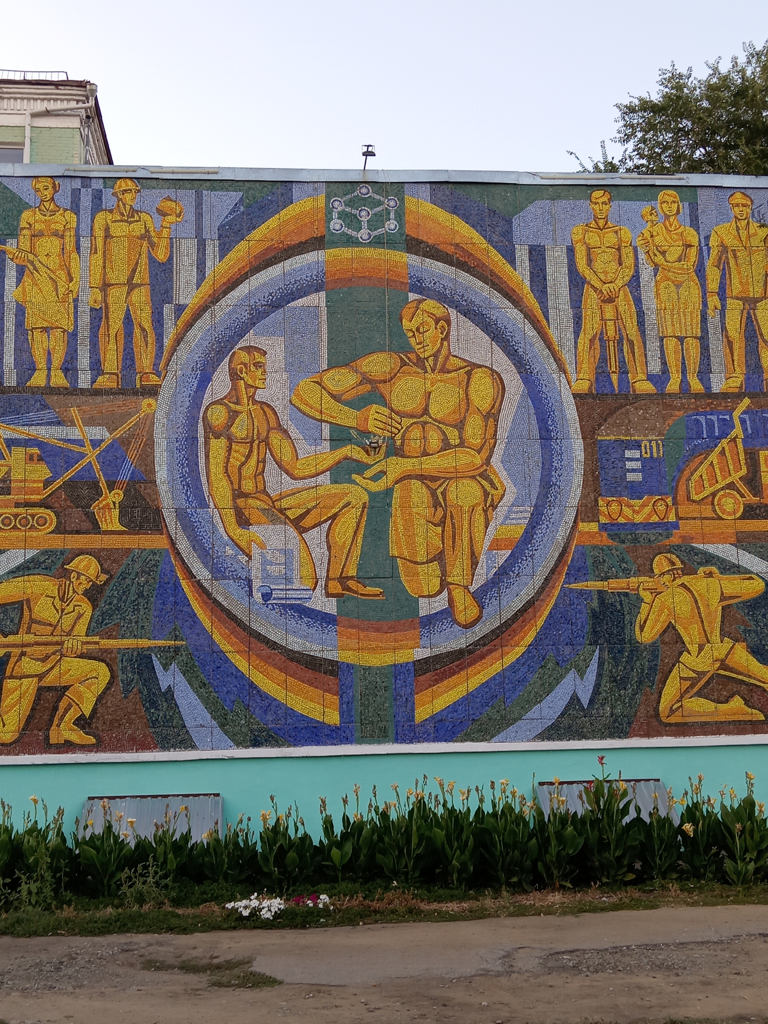 The image depicts a large, colorful mosaic mural on the exterior wall of a building, likely in the city of Rudny, Kazakhstan. The mural is comprised of many small, square tiles and features stylized figures in a socialist realist style, common in Soviet-era art.
The central part of the mural shows two muscular male figures. One is seated, holding a small object, while the other stands over him, seemingly presenting or examining it. They are surrounded by a circular motif with a rainbow-like arc.
Flanking the central figures are groups of other stylized individuals, also rendered in yellow and orange hues against a background of blues and greens. To the left, there are figures holding tools and construction equipment, including a crane and a truck. To the right, there are figures with what appear to be rifles or tools, suggesting themes of labor and possibly defense or industry. Above the figures, a molecular structure or scientific diagram is depicted in white and green tiles.
The mural covers a significant portion of the building's facade. Below the mural is a light blue band, and beneath that, a strip of green plants, including tall plants with yellow flowers and smaller white flowers in the foreground. A weathered metal panel is visible behind the plants. The ground in front of the plants is a dirt and gravel path. The sky above is clear and light-colored, suggesting it is either early morning or late afternoon. The overall impression is one of a public art installation celebrating labor and industry.