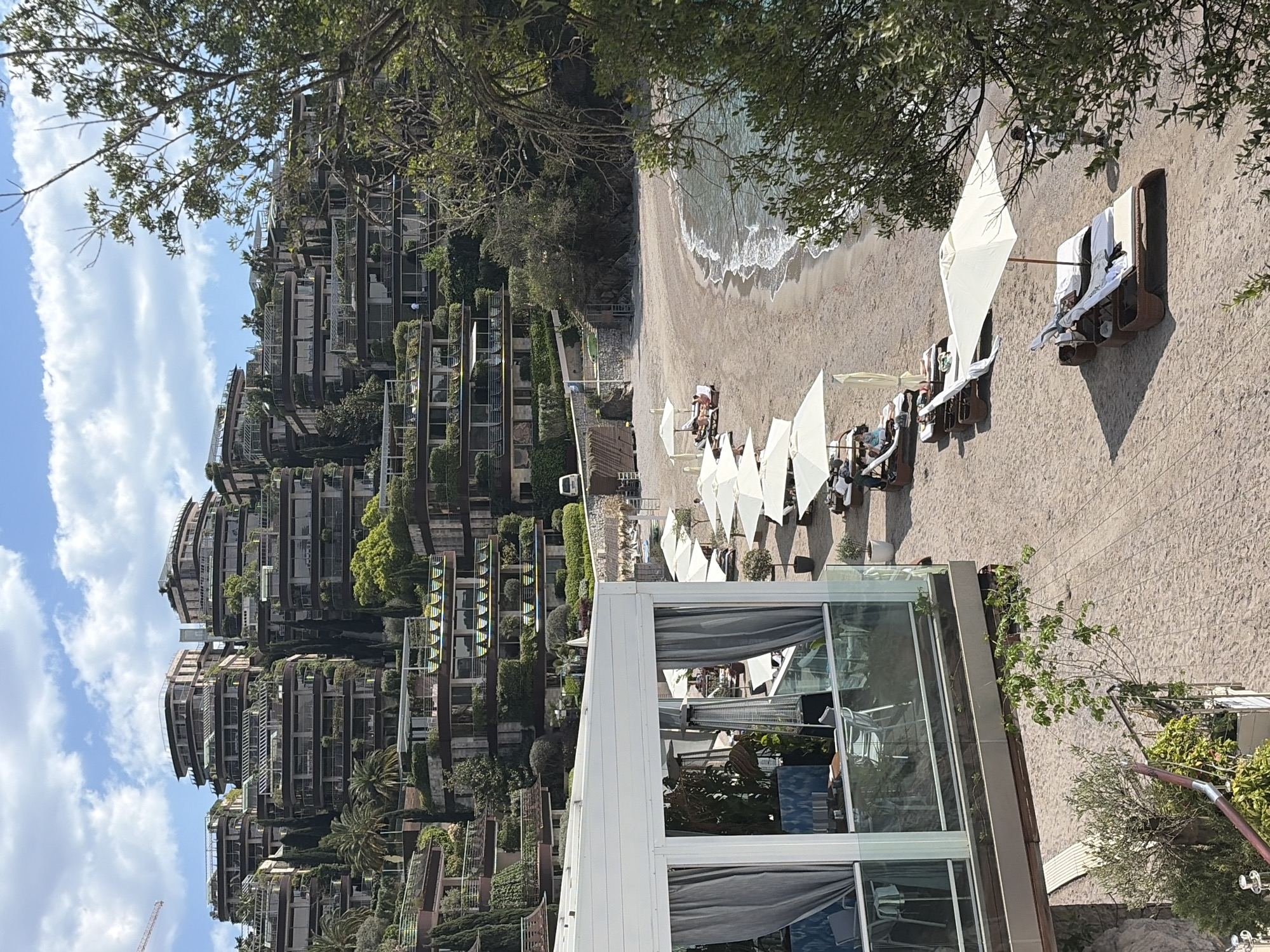 The image depicts a beach scene in Budva, Montenegro. A beach is visible, with waves coming onto the sand. Several rows of white beach umbrellas and lounge chairs are set up on the sand, and people appear to be relaxing on the beach. In the background, a building complex is built into the hillside, covered in greenery. A modern, glass-walled structure is positioned in the foreground, potentially a restaurant or bar. The sky is blue with scattered clouds, and the surrounding trees provide shade to the area.