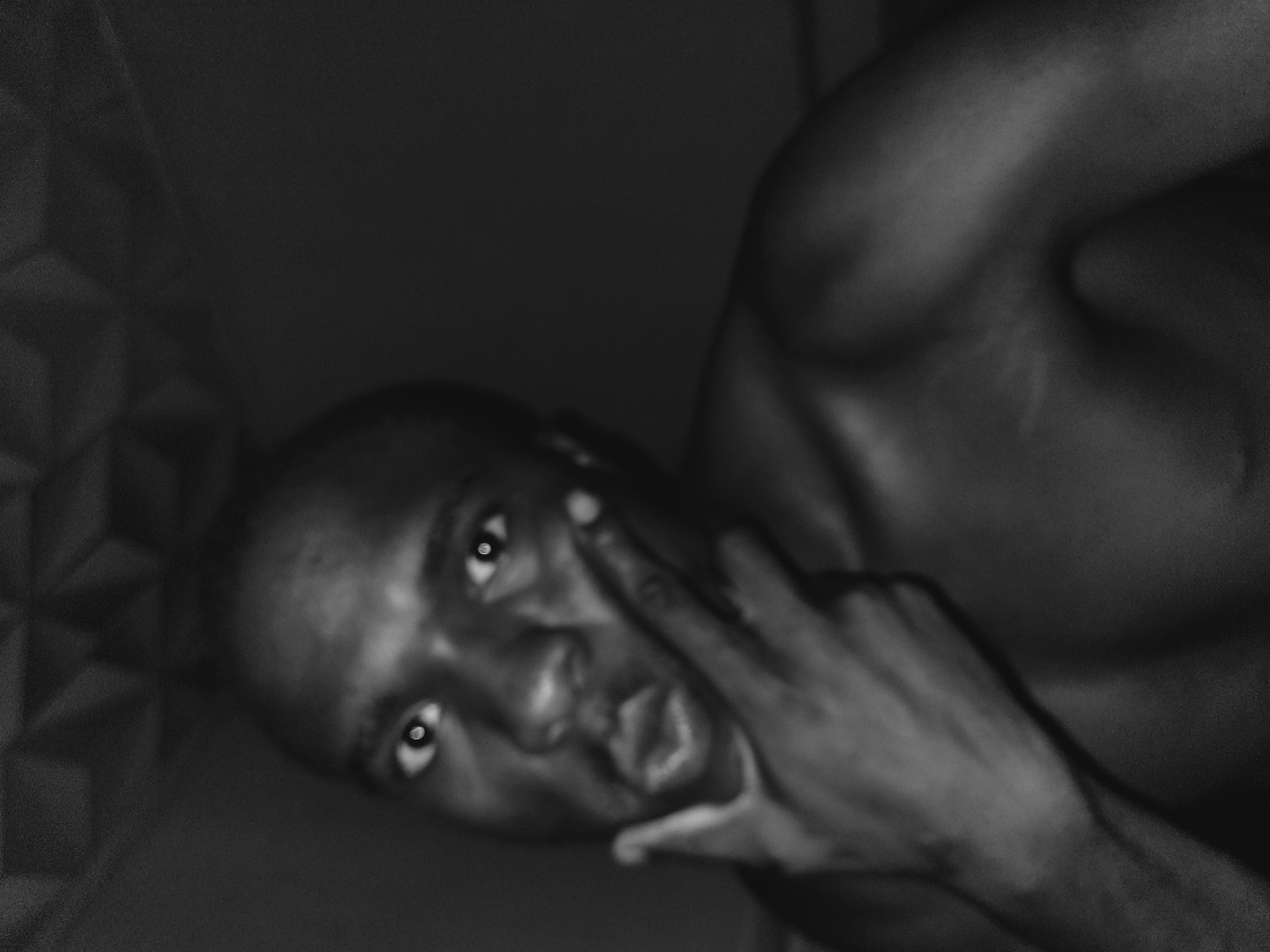 The black and white, medium-shot, close-up image depicts a shirtless young Black man in a dimly lit indoor environment. He is looking directly at the camera with his mouth slightly open and his lips puckered. His left hand is raised, with two fingers touching his lips, and his right arm is resting on his chest. The background is dark and appears to be a ceiling with a geometric pattern. There are no discernible objects or other people in the frame. The image quality is grainy, and the lighting is low, creating a mood of intimacy or mystery. It is not possible to determine the specific location in the city of Bukuru, Nigeria based on the provided image.
