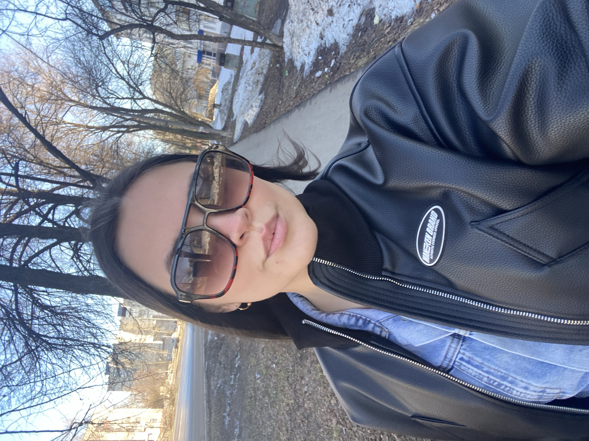 A woman is taking a selfie outdoors, seemingly on a sidewalk in Chaikovsky, Russia. She is wearing large sunglasses, a black faux leather jacket with a "Mickey Brand Authentic Overwear Apparel" patch on the right side, and a denim shirt visible underneath the jacket. The background includes bare trees, a street, and buildings with some snow patches.