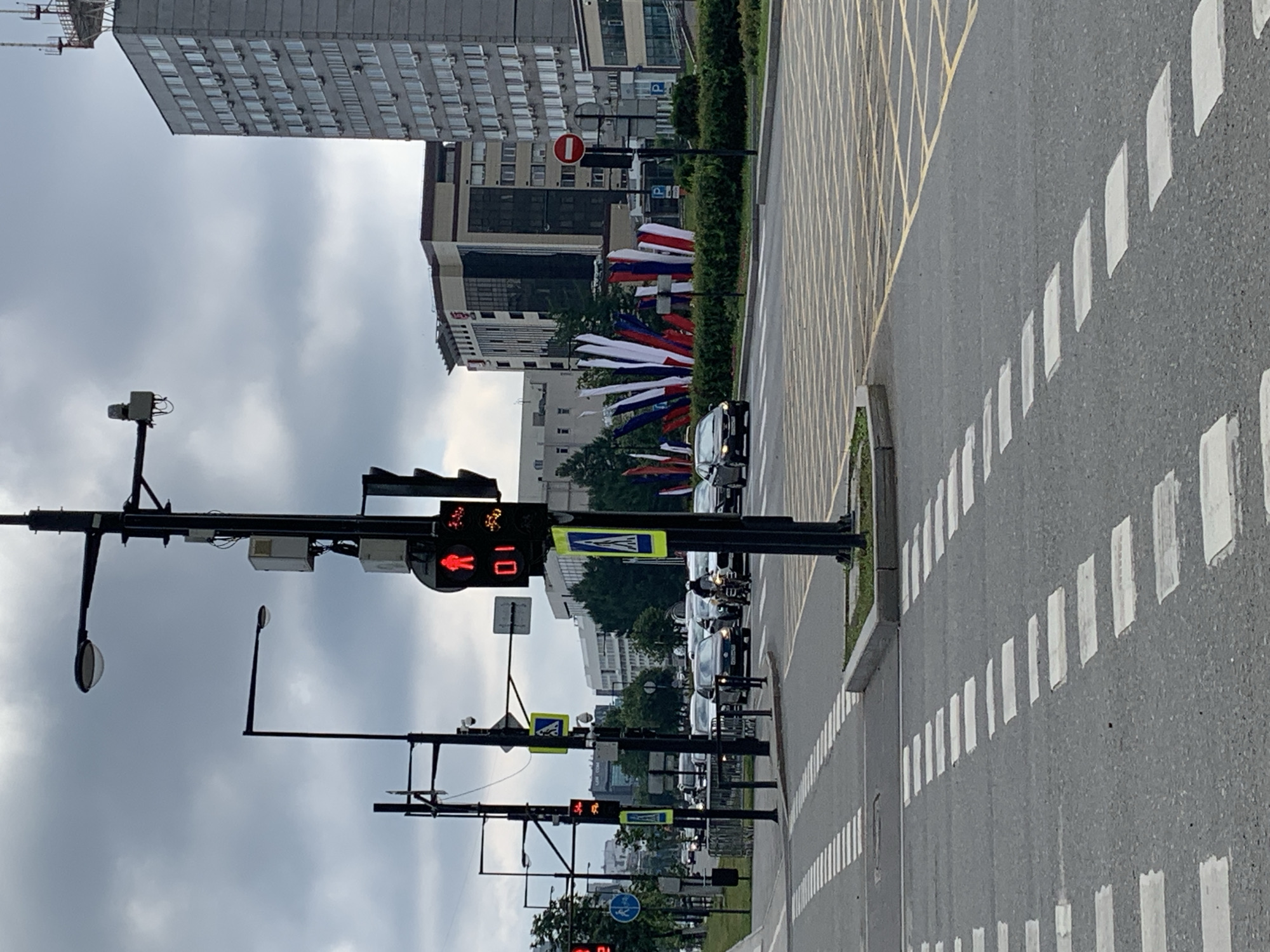 This image depicts a city street scene in Perm, Russia, likely on a cloudy day. The foreground features a pedestrian crossing with distinct white stripes painted on the asphalt. In the middle ground, traffic lights are visible, one showing a red pedestrian signal with the numeral "01". Cars and a motorcycle are stopped at the intersection. Behind the intersection, several tall buildings line the street, with one prominent grey building appearing to be a residential or office complex. A row of large flags in red, white, and blue is displayed along a manicured green hedge. The sky is overcast with grey clouds. The scene suggests a typical urban environment with traffic control systems and architectural structures.