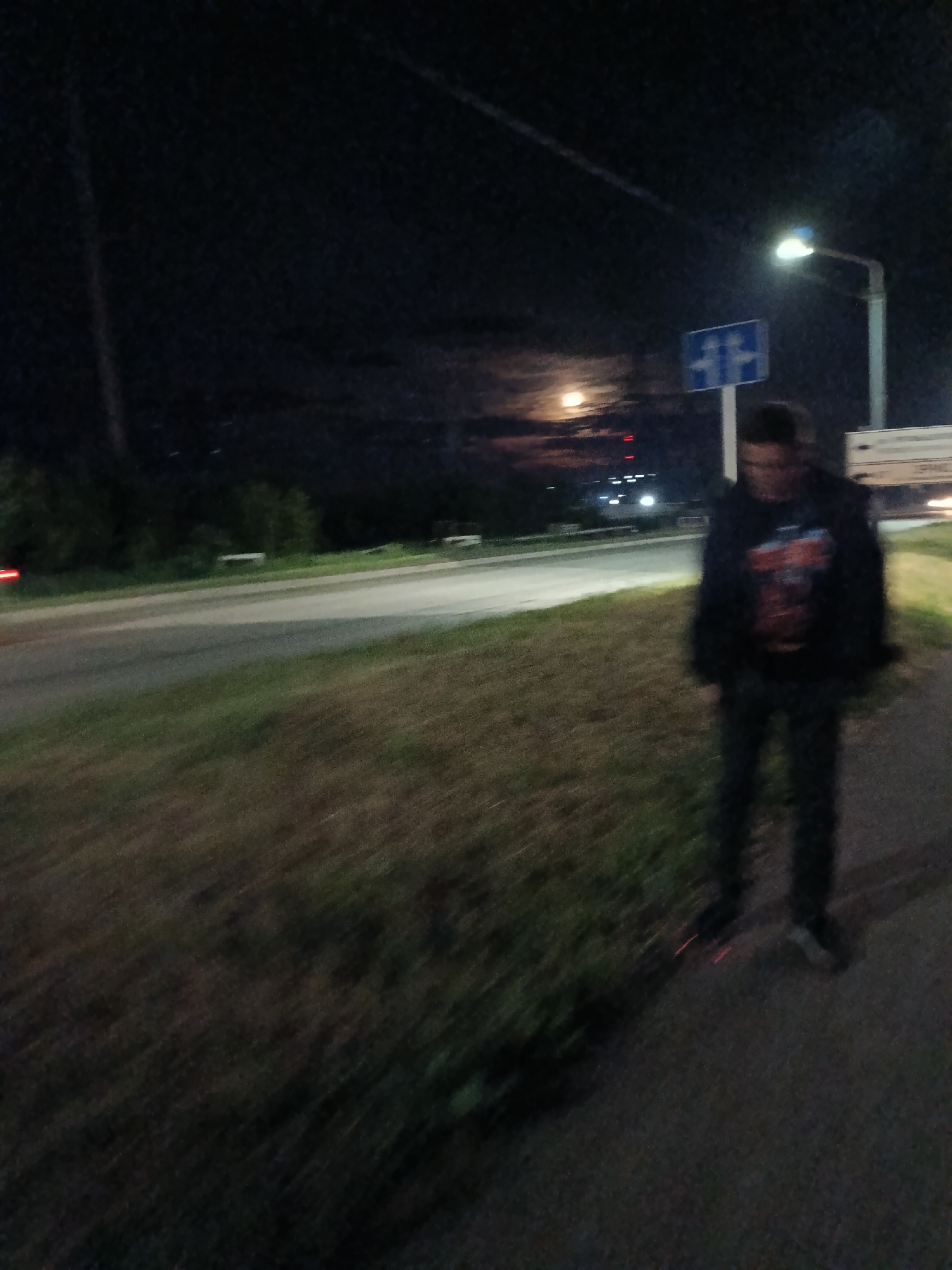 At night in Chaikovsky, Russia, a person stands on a paved sidewalk next to a grassy area and a road. The person is wearing dark clothing, including a hoodie. A road sign is visible behind the person, illuminated by a street light. Further in the background, a moon is visible in the sky. There are other lights in the distance, possibly from buildings or vehicles.