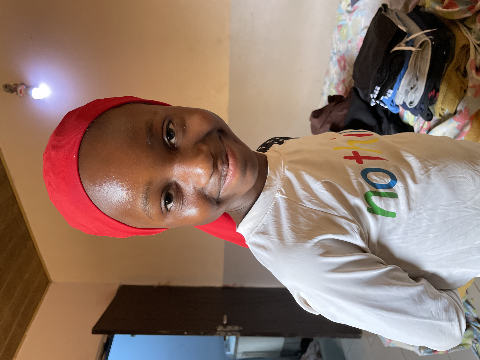 A young woman is captured in a close-up portrait, smiling brightly and looking directly at the camera. She has dark skin and is wearing a vivid red head covering, possibly a hijab or headwrap. Her attire includes a white long-sleeved shirt with colorful text across the chest. The visible letters of the text are red 'T', yellow 'O', green 'U', and a blue letter, likely spelling "INTO YOU". A natural sheen is visible on her forehead and cheeks.
The scene takes place indoors, likely within a room in a home. The background features light-colored walls and a dark brown door to the lower left. A bare, bright light bulb is prominently visible on the ceiling in the upper left portion of the image, acting as the main light source. To the right of the frame, behind the woman, a pile of various clothes (including black, blue, grey, and yellow garments) is stacked on a surface, which appears to be covered by a light-colored, floral-patterned fabric, possibly a bedspread. The overall impression is one of warmth and personal connection, consistent with a casual domestic setting in Jos, Nigeria, during evening or night due to the artificial lighting.