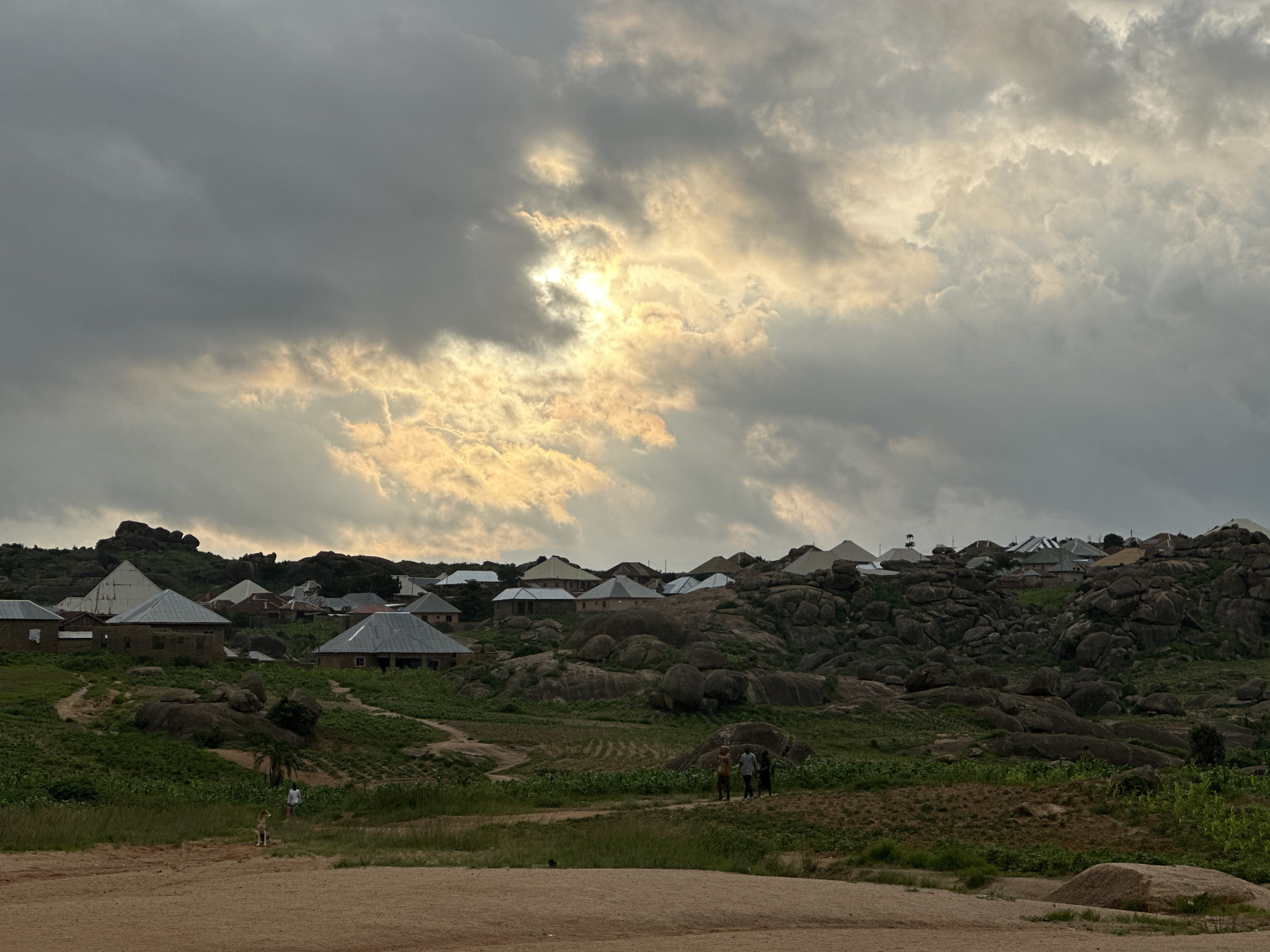 The image depicts a rural village situated on a rocky hillside, likely in the city of Jos, Nigeria. The sky is overcast with dramatic clouds, through which sunlight is breaking, casting a warm glow on the scene. The village itself consists of several buildings with corrugated metal roofs, clustered amongst large rock formations. Lush green vegetation, including crops and trees, covers the lower slopes of the hill. In the foreground, there is a sandy or dirt path. A few people are visible, including three individuals standing together in the middle ground, and one person walking in the distance near a dog. The overall atmosphere is serene and rustic, with the natural landscape dominating the scene.