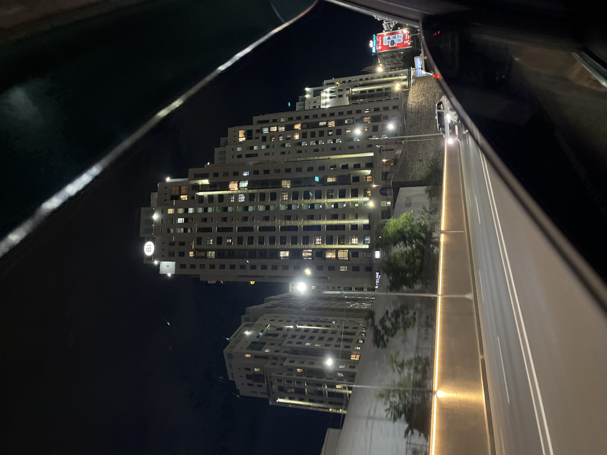 The image shows a night view of a multi-lane road in Almaty, Kazakhstan, with several tall, modern buildings on the right side of the road. The buildings are illuminated, with many windows lit up. One of the buildings has a circular logo with "BI" inside at the top. A billboard is visible on the far right. A white car is driving on the road towards the viewer. The perspective suggests the photo was taken from inside a vehicle, looking out through the window. The road has white lane markings and is bordered by a retaining wall with an illuminated strip. Trees are visible in front of the retaining wall. The sky is dark and clear.