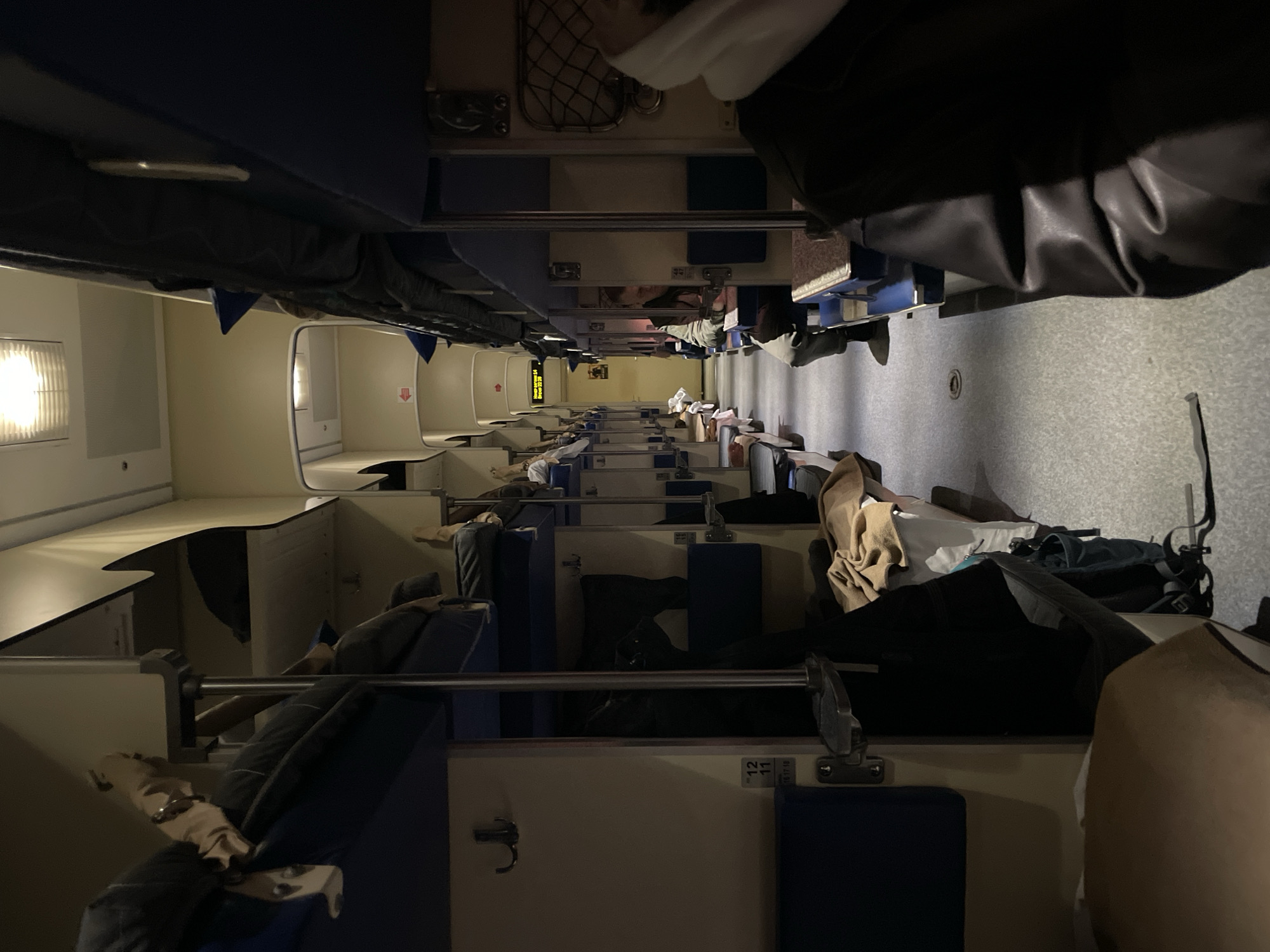 The image depicts the interior of a train car, likely a sleeper car. The space is divided into compartments with bunk beds on both sides of a narrow aisle. People are visible in some of the bunks, either sleeping or resting. Personal belongings like luggage and clothing are scattered throughout the compartments. A digital display with the time is visible in the center of the image, further down the aisle. The overall lighting is dim and possibly fluorescent.