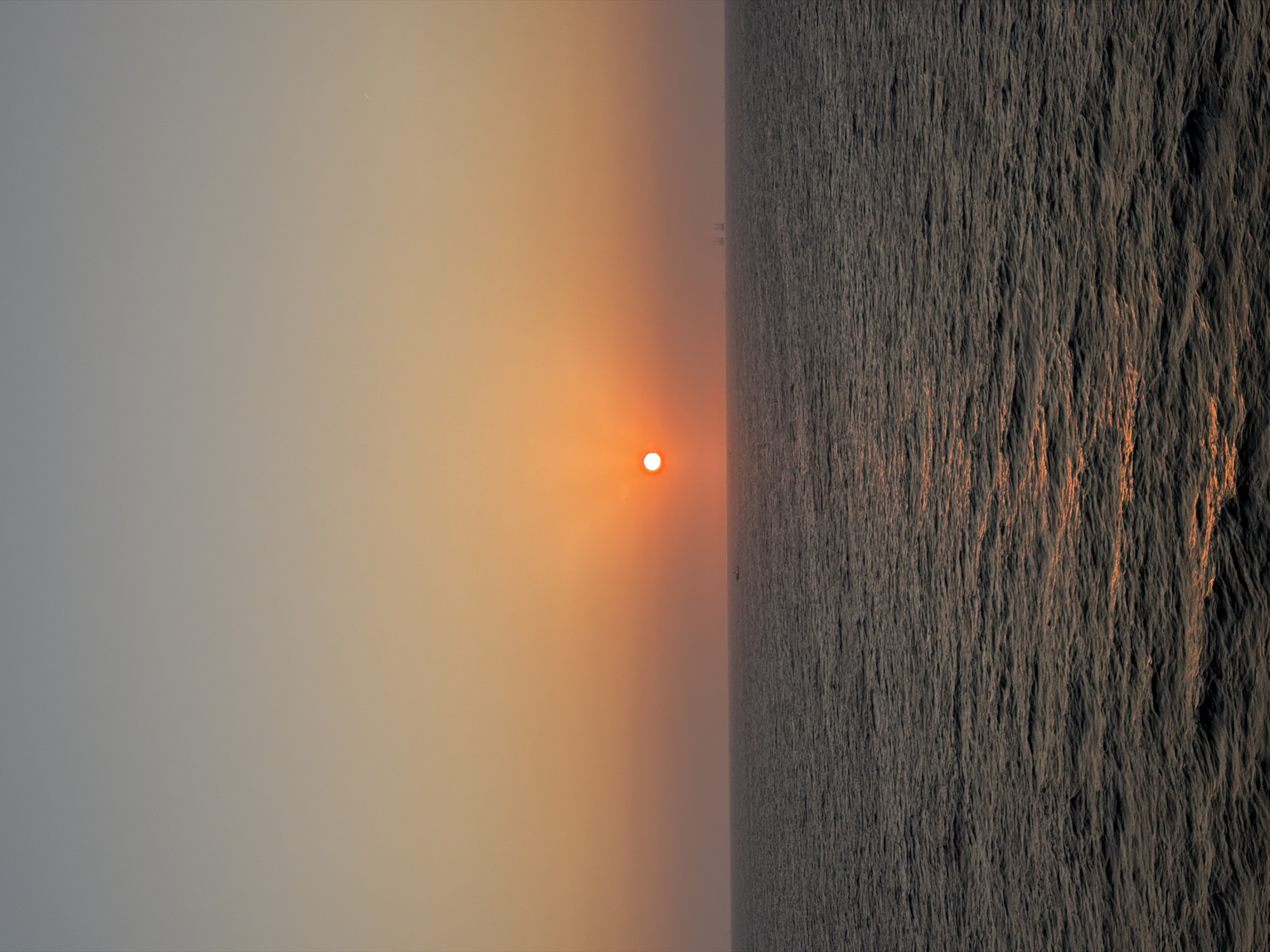 The image depicts a serene sunset or sunrise over a vast body of water, likely an ocean or large sea. The photograph is oriented vertically, but viewing it horizontally reveals a classic seascape divided by a clear horizon line.
The dominant subject is the sun, a small, intensely bright orb positioned low on the horizon, slightly to the left of the frame's center. Its core appears white-hot, surrounded by a soft, radiant orange glow that permeates the surrounding atmosphere.
The sky occupies the upper portion of the scene. It presents a smooth gradient of colors, transitioning from a cooler, muted grey-blue in the upper-left corner to warmer tones of soft orange and pink as it approaches the sun and the horizon. A tiny, almost imperceptible white speck, possibly a distant star or aircraft, is visible in the upper-left sky.
The lower half of the image is dominated by the water. Its surface is textured with countless small ripples and waves, catching the light and creating a sense of depth. While the majority of the water appears dark and slightly desaturated, the direct path of the sun's rays creates streaks of warm orange and golden reflections across its surface, particularly closer to the horizon. In the far distance, extremely faint and indistinct silhouettes, possibly of distant ships or land, can be discerned along the horizon line.
The time of day is clearly either sunset or sunrise, indicated by the low position of the sun and the characteristic warm, diffused lighting. The overall atmosphere appears slightly hazy, contributing to the soft glow around the sun and obscuring distant details. There is no visible text in the image.