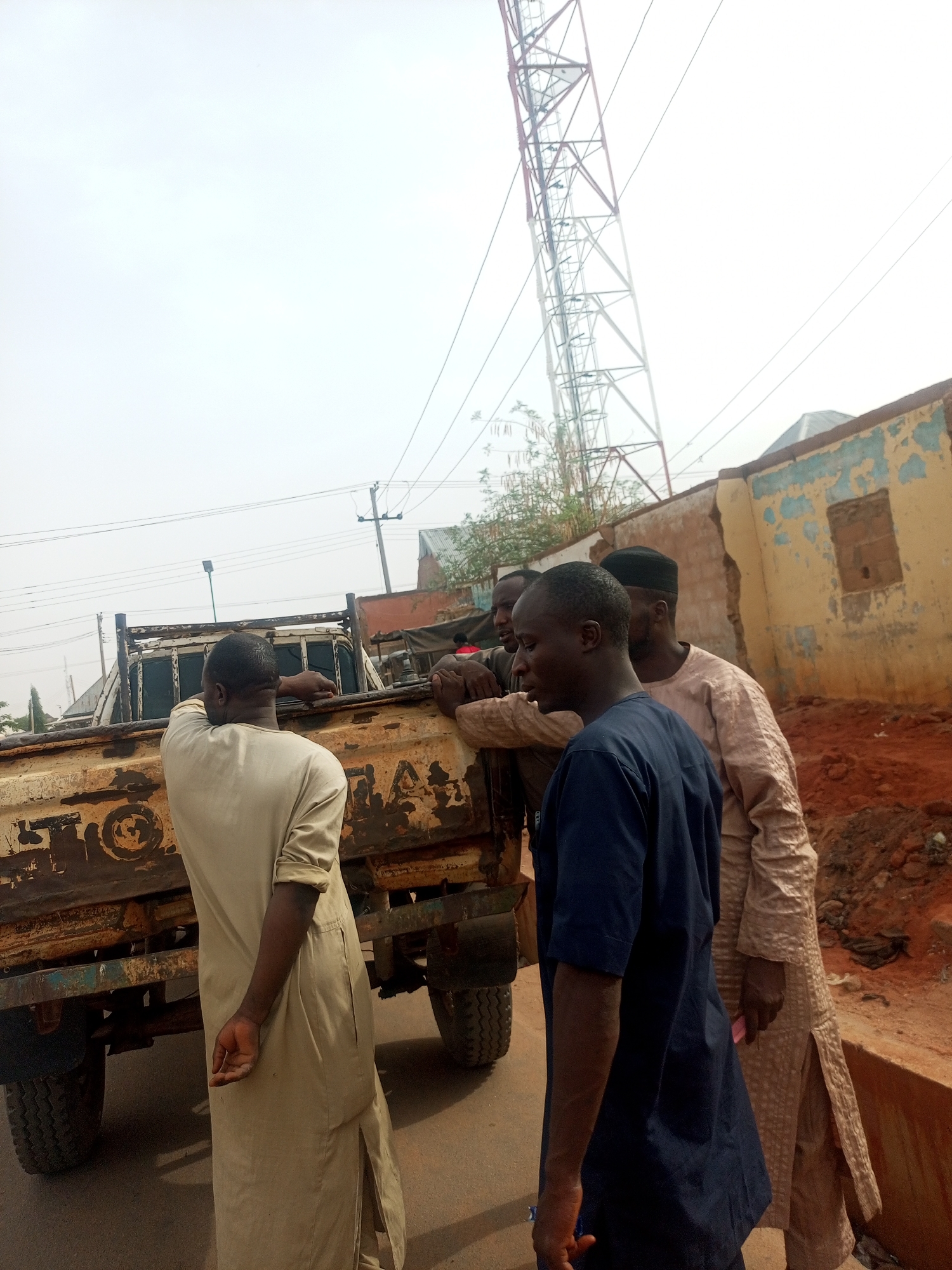 The image depicts an outdoor scene in what appears to be an urban or semi-urban environment, consistent with Gombe, Nigeria, under an overcast sky during daylight hours. Four men are gathered around a rusty utility truck.
In the foreground, a man in a light-colored, long traditional garment (possibly a kaftan) stands with his back to the camera, leaning on the heavily rusted tailgate of a pick-up truck. Faint, faded lettering "TOYOTA" is partially visible on the truck's rear. Beside him, another man in a dark blue short-sleeved garment faces right, looking off-camera. Behind these two, two other men are partially visible; one wears a light brown patterned long garment and a dark cap, looking towards the man in blue, while the fourth man is mostly obscured. Their attire suggests local traditional clothing.
The truck exhibits significant wear and rust, indicating its age and heavy use. In the background, a tall, red and white metal lattice tower, likely a telecommunications or electricity mast, extends upwards against the pale sky. Overhead, utility lines are strung across the frame. To the right, a dilapidated building or wall with peeling light blue paint revealing a yellow or tan base and exposed reddish-brown earth or brickwork is visible. The ground in the vicinity is reddish-brown soil, suggesting unpaved areas or recent disturbance.
The men appear to be engaged in a casual gathering or conversation. No specific event is clearly identifiable beyond this interaction. The scene portrays an everyday moment in a working or developing setting.
