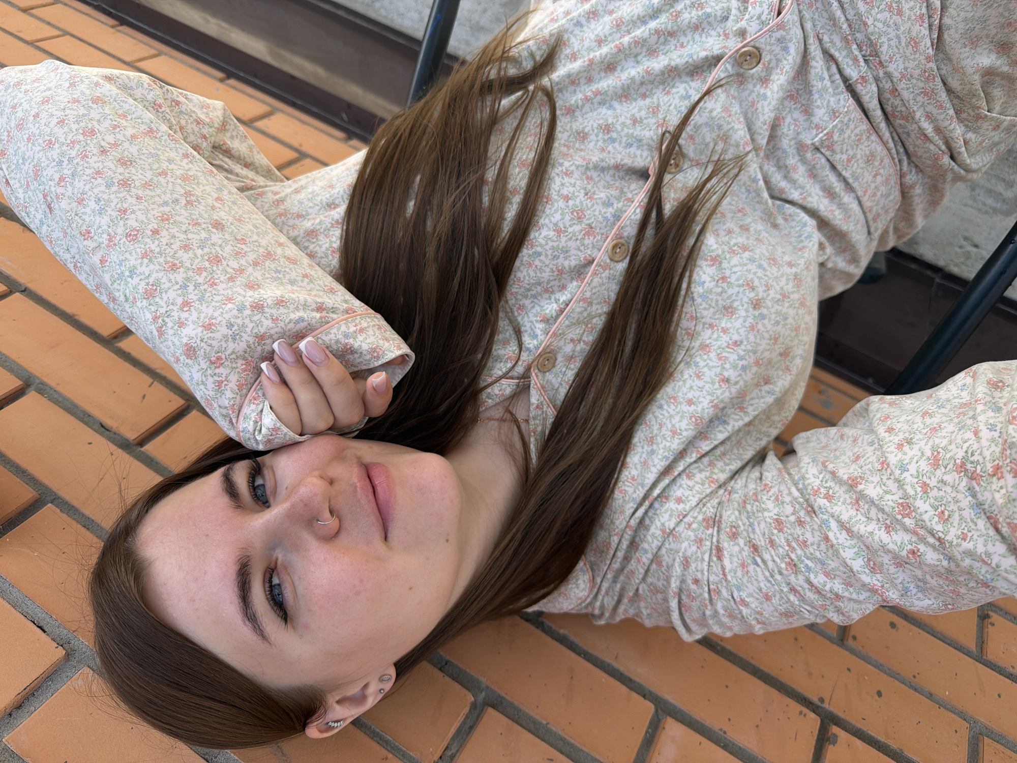 A young woman with brown hair and blue eyes is lying down on a brick wall. She is wearing a floral pajama set. Her right arm is bent and resting on her head. Her left hand is holding a cup. The background is a brick wall and a blurred background of what appears to be a city. The location is in Chaikovsky, Russia.