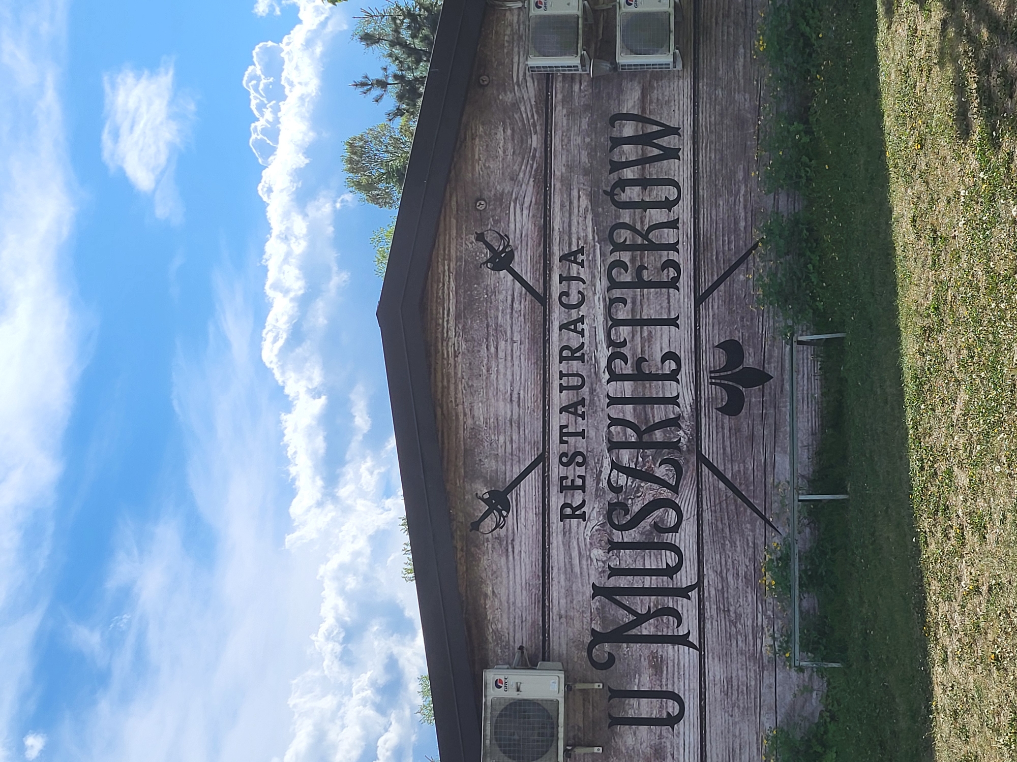 A daytime shot shows a wooden sign for "Restauracja U Muszkieterow" (Musketeers Restaurant) in Unknown, Unknown. The sign is adorned with musketeer sword designs and a fleur-de-lis. There are air conditioning units affixed to the sign, and it stands adjacent to a grassy area. The background features a bright blue sky with white clouds. The sign is captured in a slightly angled orientation.