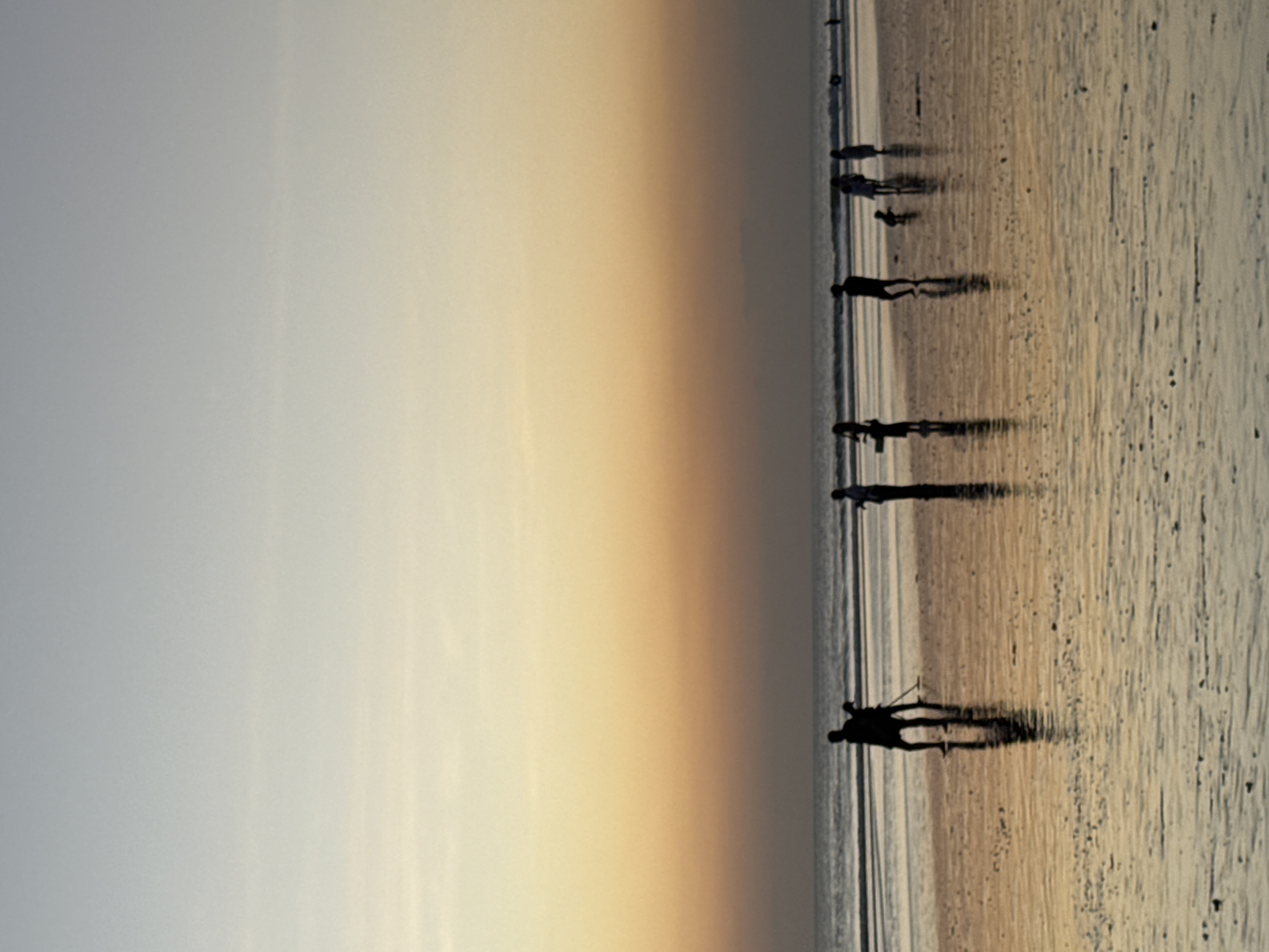 The image depicts a beach scene in Kuta, Indonesia, during what appears to be sunset. The sky displays a gradient of colors, transitioning from pale blue at the top to a warm orange and yellow closer to the horizon. The ocean is calm, with gentle waves lapping at the shore.
Several individuals are visible as silhouettes on the wet sand. In the foreground and slightly to the left, two figures stand, one with what could be a tripod or a long object. Further to the right, a group of people are spread out along the beach, some walking, others appearing to be standing or interacting. One small figure, potentially a child, is closer to the ground in the middle of the group. The wet sand reflects the colors of the sky, creating a shimmering surface. The overall atmosphere is serene and peaceful.