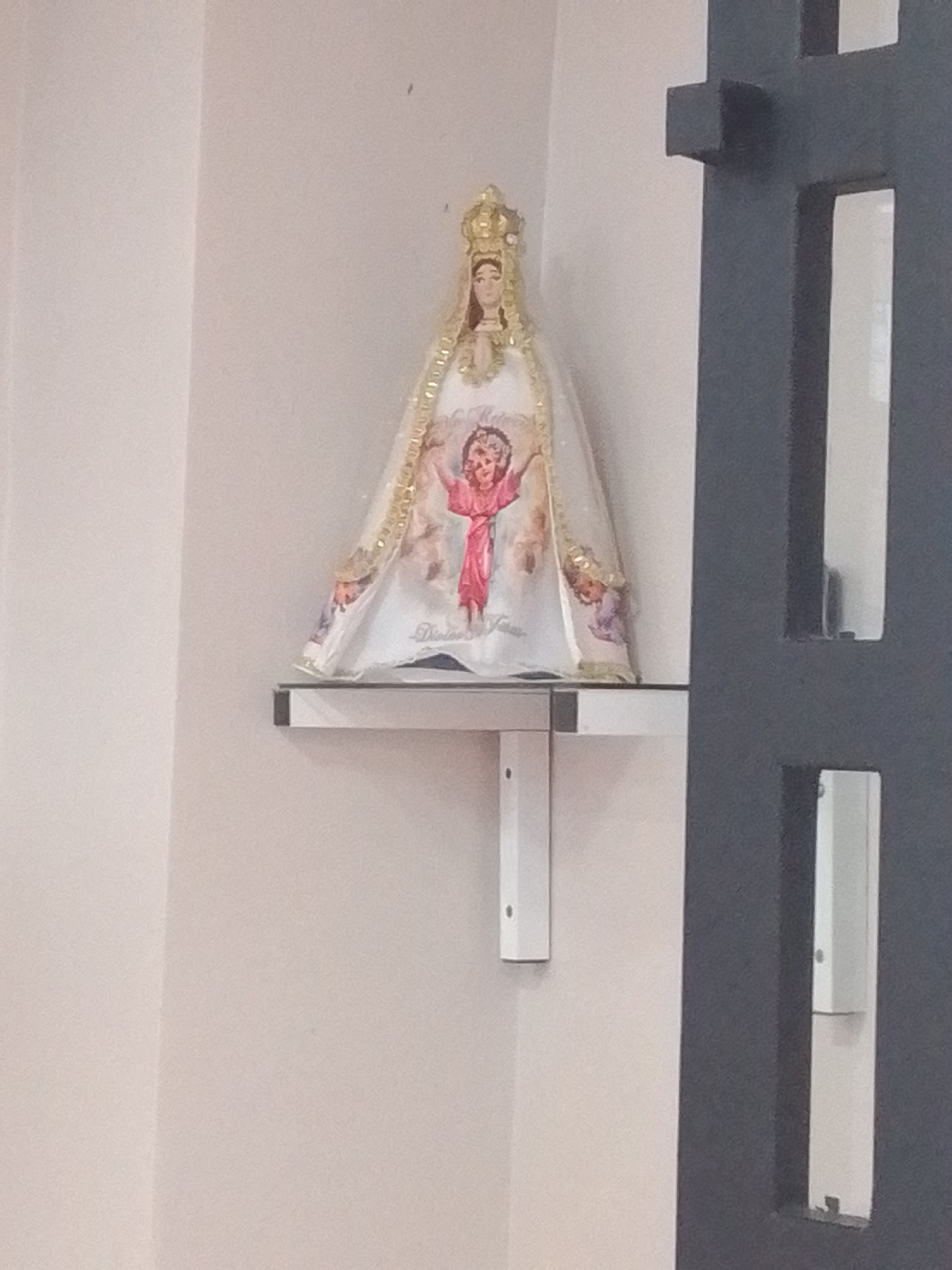 The image shows a statue of the Virgin Mary holding the infant Jesus, placed on a shelf mounted on a light-colored wall. The statue is adorned with a crown and a richly embroidered robe featuring gold trim. A depiction of the infant Jesus is visible on the front of the robe, with pink and purple hues. The shelf itself is made of a light material with a metallic edge and is supported by a white bracket. To the right of the shelf, a dark gray, opaque structure with rectangular cutouts is visible, suggesting it might be part of a doorway or screen. The background is a plain, light-colored wall, with a faint shadow or line indicating a corner. The lighting appears to be artificial and even, providing no indication of the time of day. The overall scene is simple and devotional, possibly situated in a religious establishment or a home. There is no visible text that can be clearly deciphered. The location context provided is Juan Griego, Venezuela.