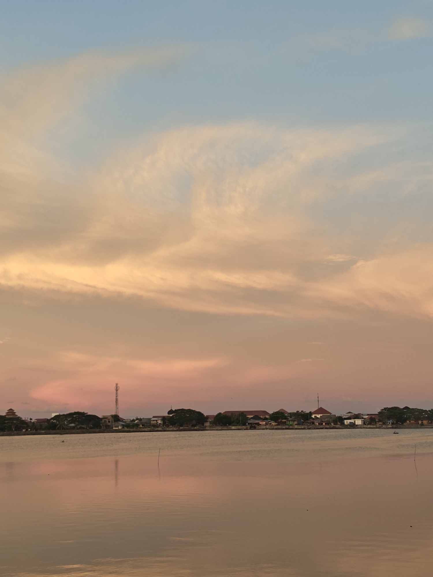 The image captures a serene sunset over the waters in Lhokseumawe, Indonesia. The sky is a canvas of soft pastel hues, transitioning from a pale blue at the top to warm oranges and pinks lower down, painted with wispy clouds. In the distance, a linear, low-rise urban landscape stretches along the horizon, characterized by residential buildings, trees, and a prominent telecommunications tower. The water in the foreground reflects the colors of the sky, creating a tranquil and symmetrical mirrored effect. A few small, dark specks in the water, likely small boats or debris, and a thin wooden pole emerging from the water add subtle details to the scene. The overall atmosphere is one of peaceful twilight in a coastal city.