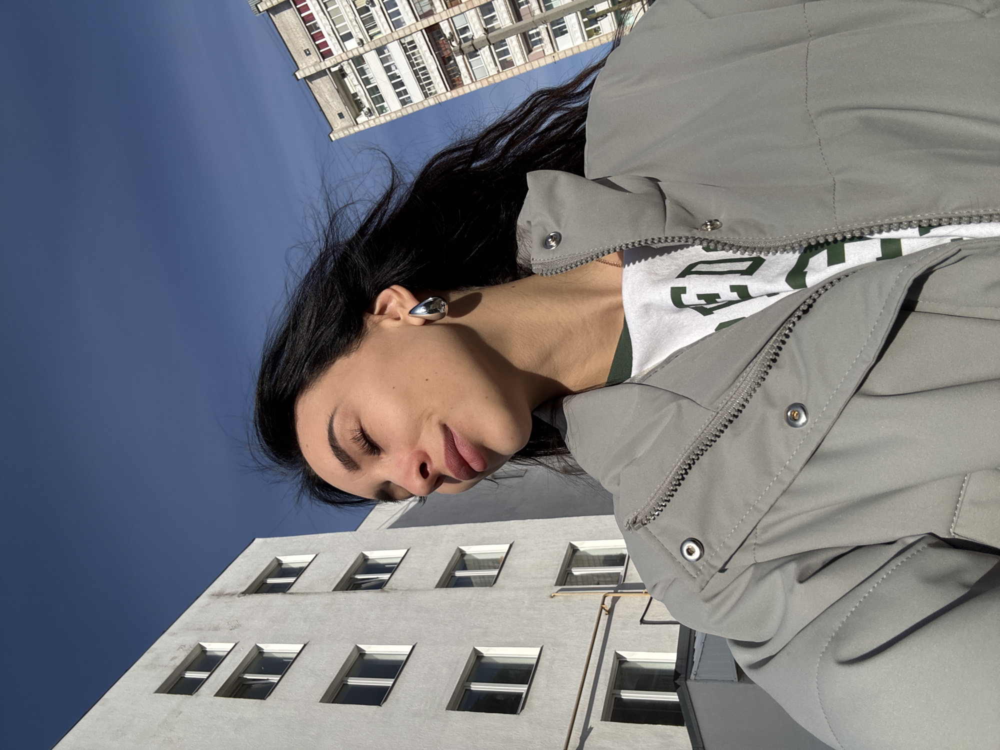 The media file shows a low-angle outdoor shot of a young woman set against a clear blue sky and urban buildings in Chaikovsky, Russia. The image is oriented in a portrait fashion, rotated 90 degrees clockwise, giving the impression of the woman lying down and looking downwards, though the building architecture suggests an upright perspective was originally intended.
The primary subject is a young woman, seen from the chest up on the left side of the frame. She has long, dark hair, light skin, and her eyes are closed or mostly closed, with a peaceful expression. Her lips are slightly parted. She is wearing a grey puffer jacket over a white top. A single, distinctive silver-toned, fluid-shaped earring is visible on her left ear. A thin, delicate chain necklace is also visible around her neck.
In the background, two light-colored multi-story buildings are visible. One occupies the upper right portion, featuring numerous windows, some with balconies, and air conditioning units, suggesting a residential or office complex. The second building is in the lower left, with several rows of rectangular windows. The sky is a vibrant, clear blue, indicating sunny weather. Bright sunlight casts distinct shadows, suggesting it's either morning or afternoon.
Visible text includes partial green block letters on her white t-shirt, beneath the open grey jacket. The letter "G" is clearly discernible, along with a portion of what appears to be another letter (possibly "A" or "E").