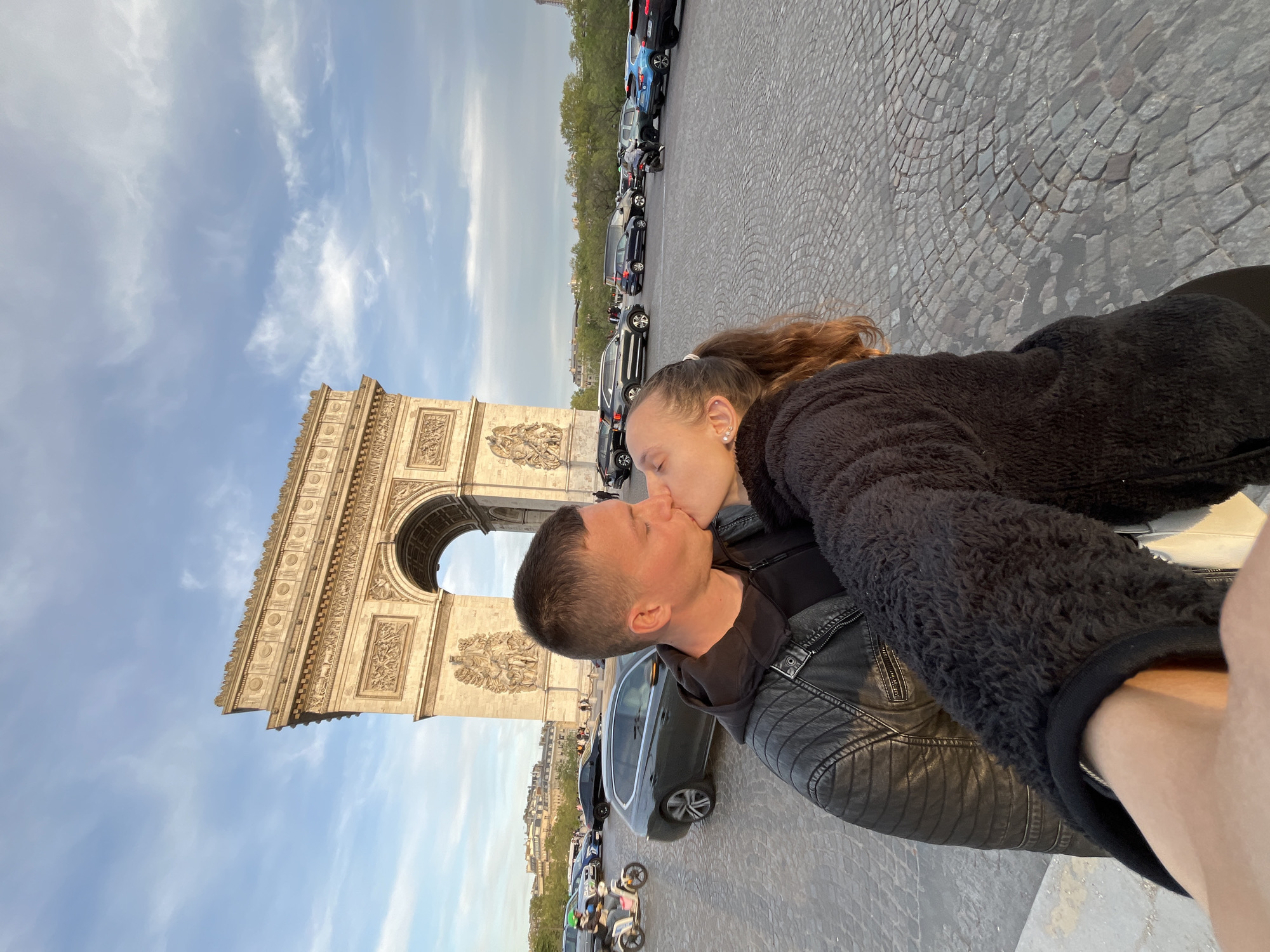 The image captures a couple kissing in Paris, France, with the iconic Arc de Triomphe serving as a prominent backdrop. In the foreground, the man, with short dark hair, is seen in profile, wearing a black leather jacket over a dark top. He is leaning in to kiss the woman, whose face is partially obscured by the embrace. She has long, reddish-brown hair tied in a ponytail and is wearing a black, fluffy jacket. The shot appears to be a selfie, indicated by a hand visible in the bottom right corner holding the camera.
The Arc de Triomphe stands to the left and behind the couple, its ornate stone facade illuminated by the warm, golden light of what appears to be late afternoon or early evening. The sky above is a clear, light blue with scattered wispy white clouds. Behind the monument and extending into the mid-ground, parked cars and motorbikes line a street, with trees and distant buildings visible beyond. The ground where the couple is standing is paved with cobblestones. The scene evokes a romantic and memorable moment in a famous location. No visible text is present in the image.