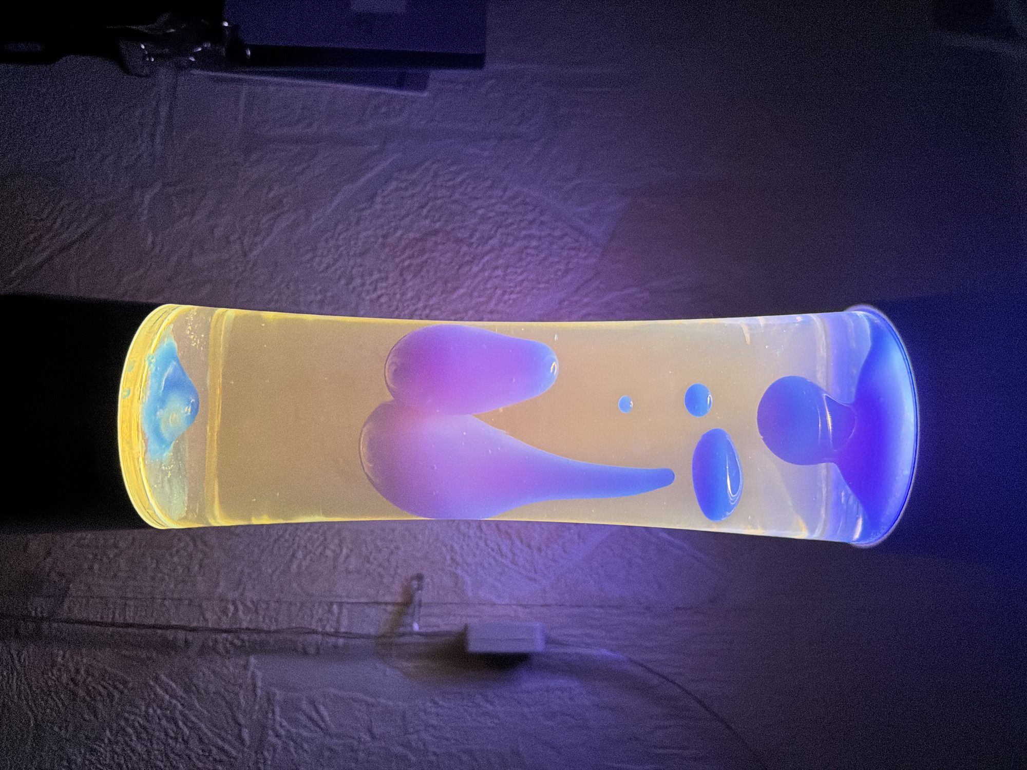 A lava lamp lies horizontally on a textured surface. The liquid inside is yellow, and blue/purple blobs float within. A power cord with a boxy adapter is visible below the lamp. The background includes textured surfaces, an undefined darker area, and an unidentified reflective rectangular object. There is no evidence in the image to support the location being in Izhevsk, Russia.