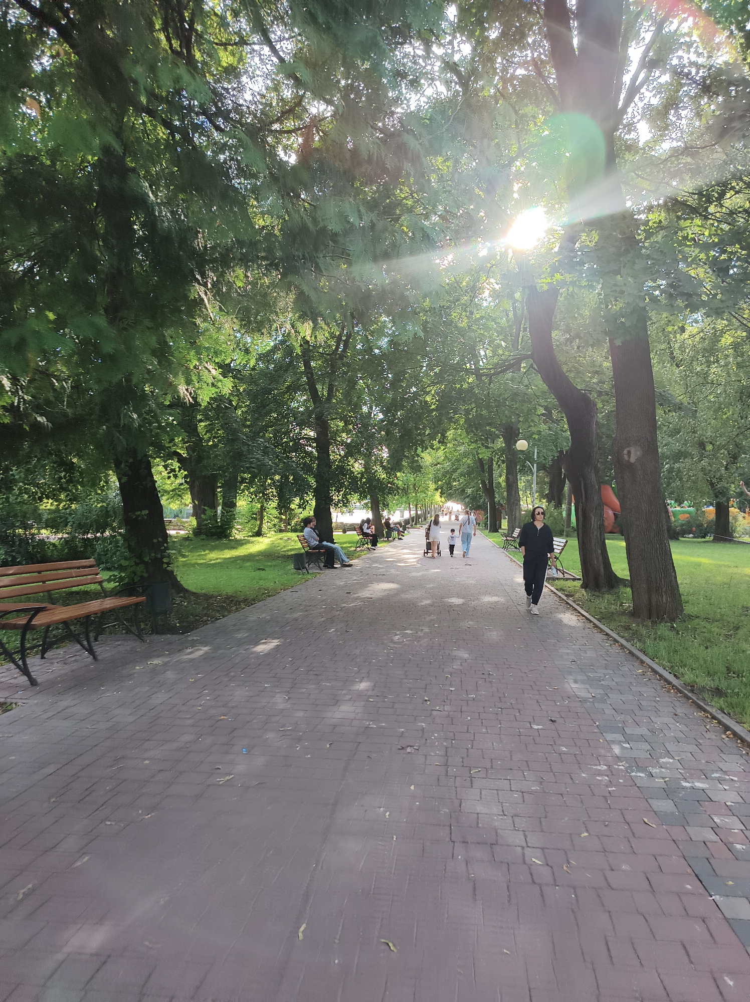 The image depicts a paved pathway in a park in Ternopil, Ukraine. The pathway is lined with mature trees on both sides, casting shadows on the ground. Sunlight filters through the leaves of the trees, creating a dappled effect. Several people are visible walking along the pathway and sitting on benches. In the foreground, a wooden bench is positioned to the left of the path. Further down the path, groups of people are walking, some with children. In the background, glimpses of a playground area with colorful structures can be seen. The overall scene suggests a peaceful day in a public park.
