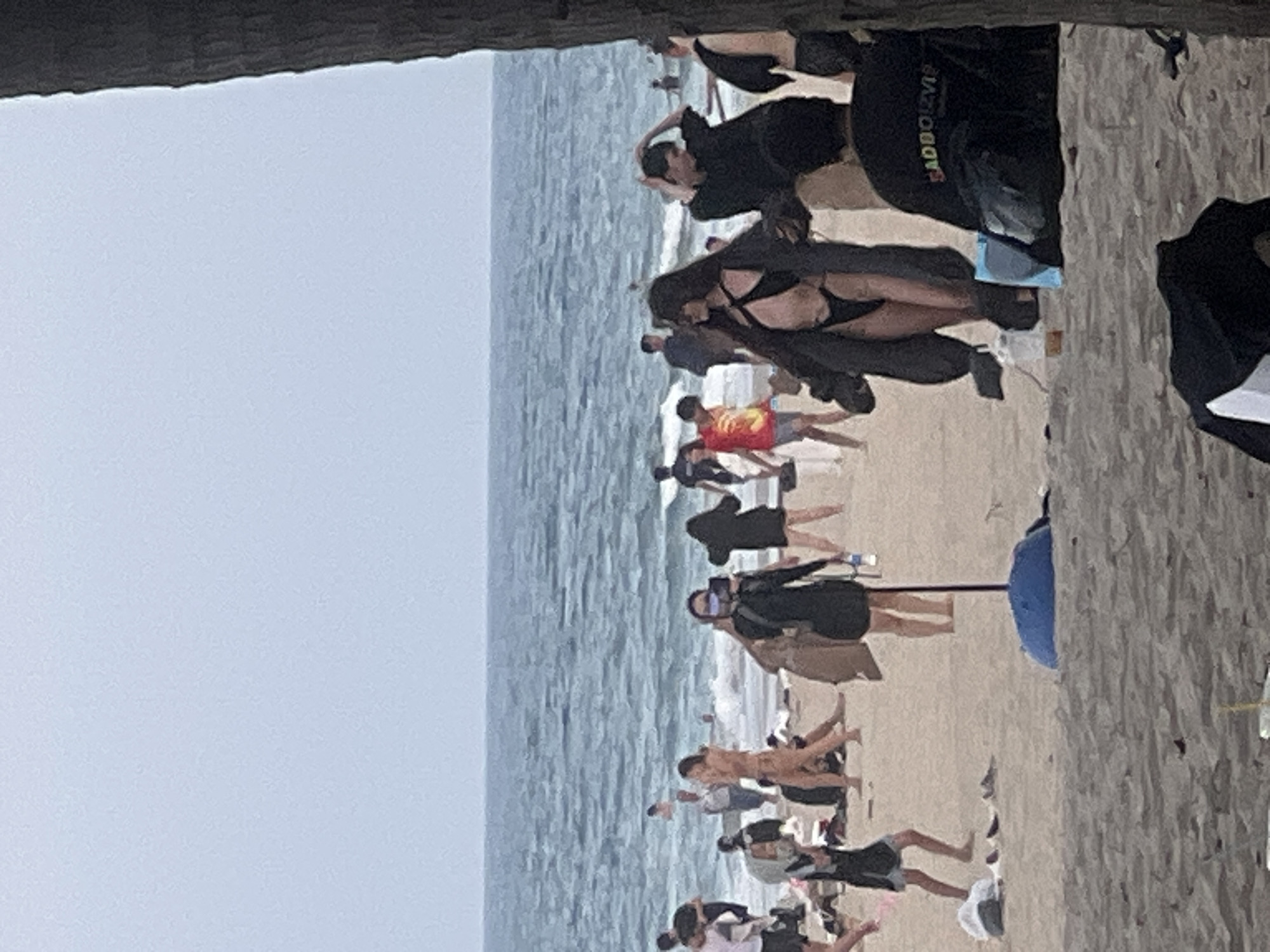 The image depicts a crowded beach scene in Da Nang, Vietnam, under an overcast sky. The foreground shows sandy beach with several people in casual attire, some sitting and others standing. A blue umbrella base is visible in the sand. In the midground, a group of people are walking and standing on the beach, some wearing swimwear. The background features the ocean with moderate waves, and several individuals are visible in the water. A person in the midground is wearing a black bikini with a dark coat over it, and another person next to them is wearing a black t-shirt. A large tree trunk is on the right side of the frame, partially obscuring the scene. The overall atmosphere appears to be one of leisure and social gathering at a coastal location.
