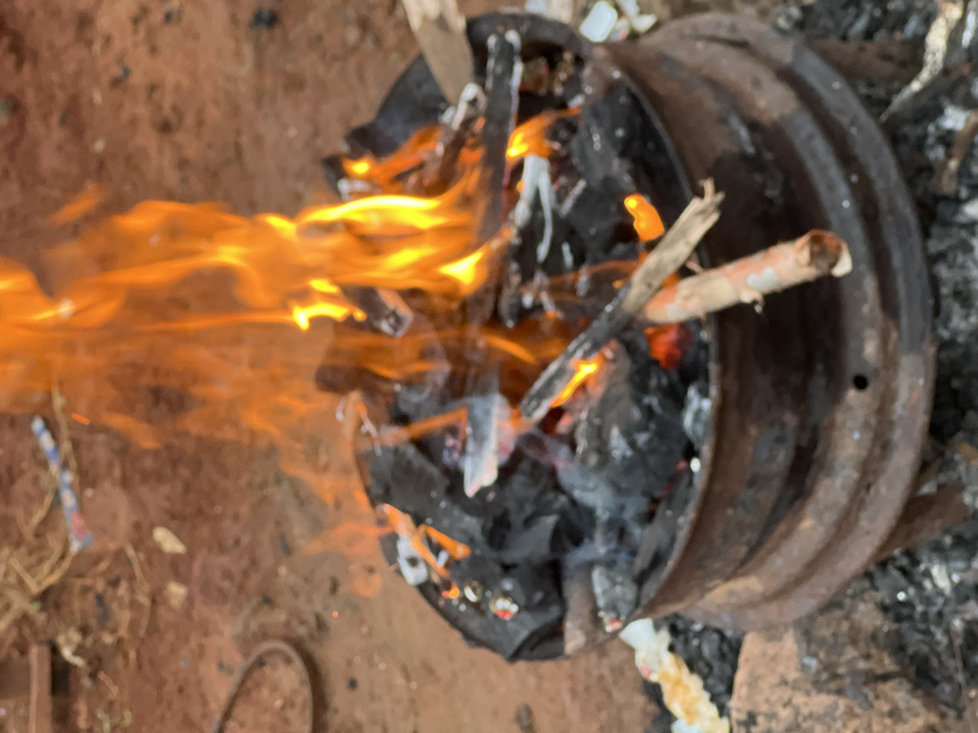 A close-up, slightly blurry shot captures a small, makeshift fire burning in a rusty metal container, possibly an old tire rim, situated on reddish-brown earth. The fire is fueled by charcoal and small pieces of wood, with vibrant orange flames leaping upwards. The background is indistinct, showing more of the earthy terrain with scattered debris. The scene appears to be an outdoor setting in the city of Nkpor, Nigeria, with the arid ground and the way the fire is contained suggesting a common practice for cooking or waste disposal in the area. There are no people visible in the image.