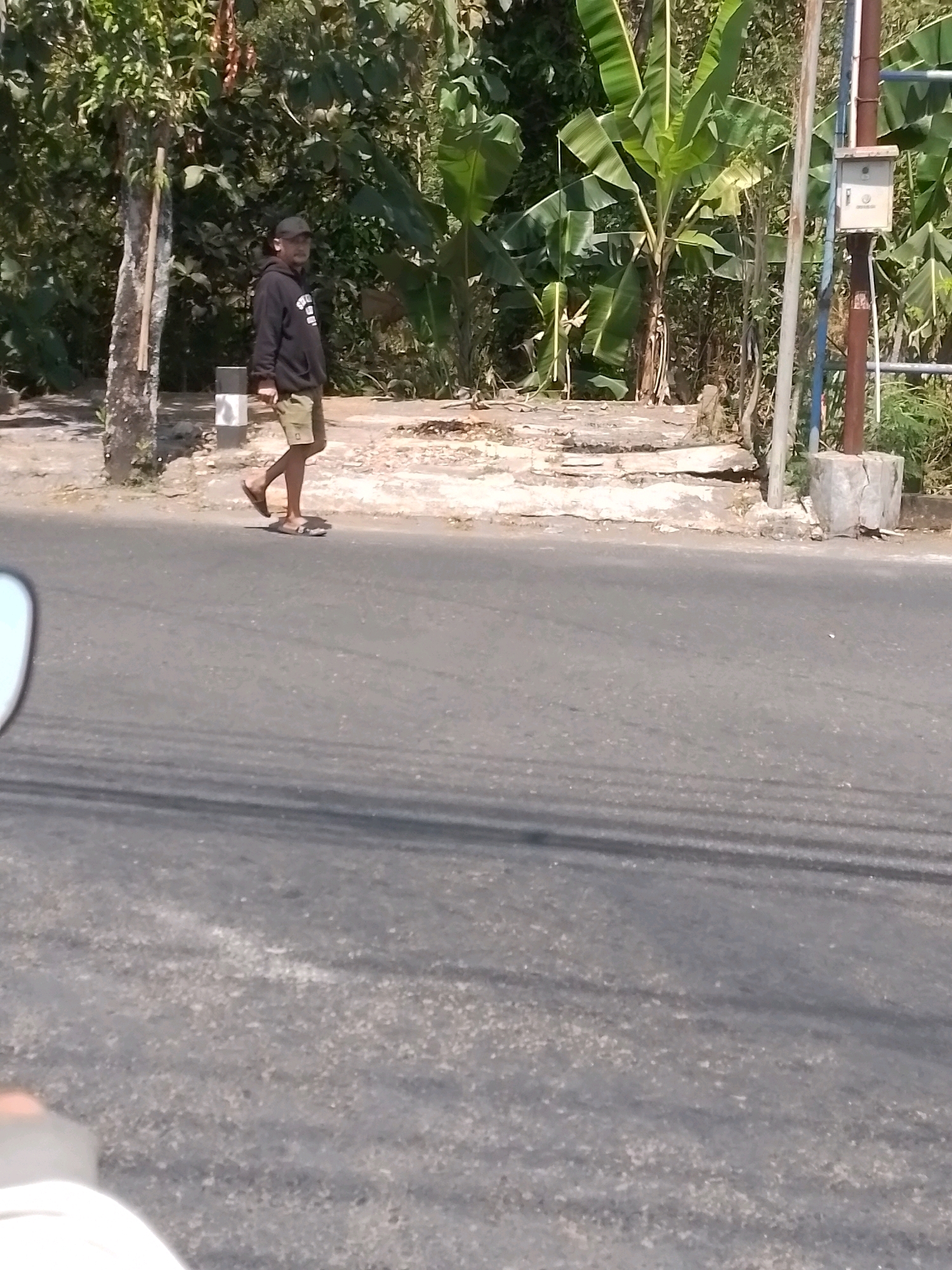 A man wearing a black hoodie, tan shorts, a baseball cap, and flip-flops walks along the side of a paved road in Wonosari, Indonesia. He is looking towards the left of the frame, with his right leg extended in a walking motion. Behind him, a narrow paved area runs alongside the road, bordered by a grassy embankment and dense vegetation including banana trees. A light pole with a small electrical box is visible on the right side of the embankment, and a thin tree stands to the left of the man. The road surface is asphalt, with faded white lines visible. The scene is illuminated by bright sunlight, casting shadows.