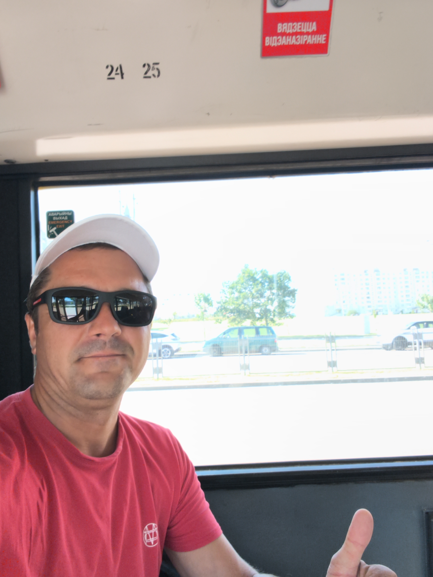 The image shows a man with sunglasses and a white cap, wearing a red t-shirt, taking a selfie inside a bus or tram. He is giving a thumbs-up gesture. Through the window, a cityscape is visible, with buildings, trees, and vehicles. A sign on the ceiling of the vehicle reads "24 25" in black lettering. Another sign in red with white text is partially visible, likely indicating safety instructions. There is also a green sticker with "EMERGENCY EXIT" and text in a Cyrillic script. Based on the Cyrillic script and the style of buildings, the location is likely Minsk, Belarus.
