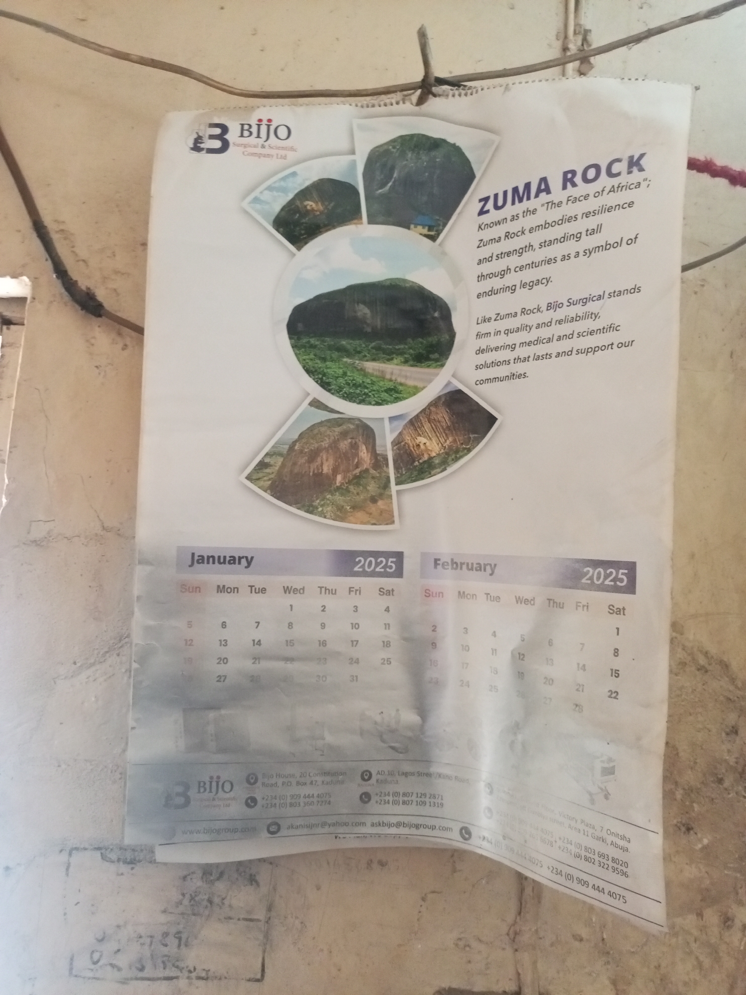 A calendar for the year 2025, specifically showing January and February, hangs on a weathered wall in what appears to be an indoor or covered outdoor setting in Maiburiji, Nigeria. The calendar is held up by a piece of string or wire attached to another wire running horizontally across the wall. The wall itself is light-colored, possibly painted, but shows signs of dirt and wear.
The calendar features a prominent image of Zuma Rock, described as "The Face of Africa," with multiple smaller images of the rock arranged around a central, larger image. Text associated with the rock highlights its "resilience and strength" as a "symbol of enduring legacy." The calendar is an advertisement for "Bijo Surgical & Scientific Company Ltd," which states it "stands firm in quality and reliability, delivering medical and scientific solutions that lasts and support our communities," likening its values to Zuma Rock.
The calendar pages clearly display the days of the week (Sun, Mon, Tue, Wed, Thu, Fri, Sat) and the dates for January and February 2025. January begins on a Wednesday (1st) and February on a Saturday (1st).
At the bottom, the calendar includes contact information for Bijo Surgical & Scientific Company Ltd, with physical addresses in Kaduna (Bijo House, 20 Constitution Road, and AD.16, Lagos Street/Kano Road), and additional offices in Abuja (Area 11 Garki) and Onitsha (Victory Plaza). Multiple Nigerian phone numbers and email addresses (akanisijnr@yahoo.com, askbijo@bijogroup.com) are listed, along with their website, www.bijogroup.com. Some illegible scribbles or markings are visible on the wall beneath the calendar.