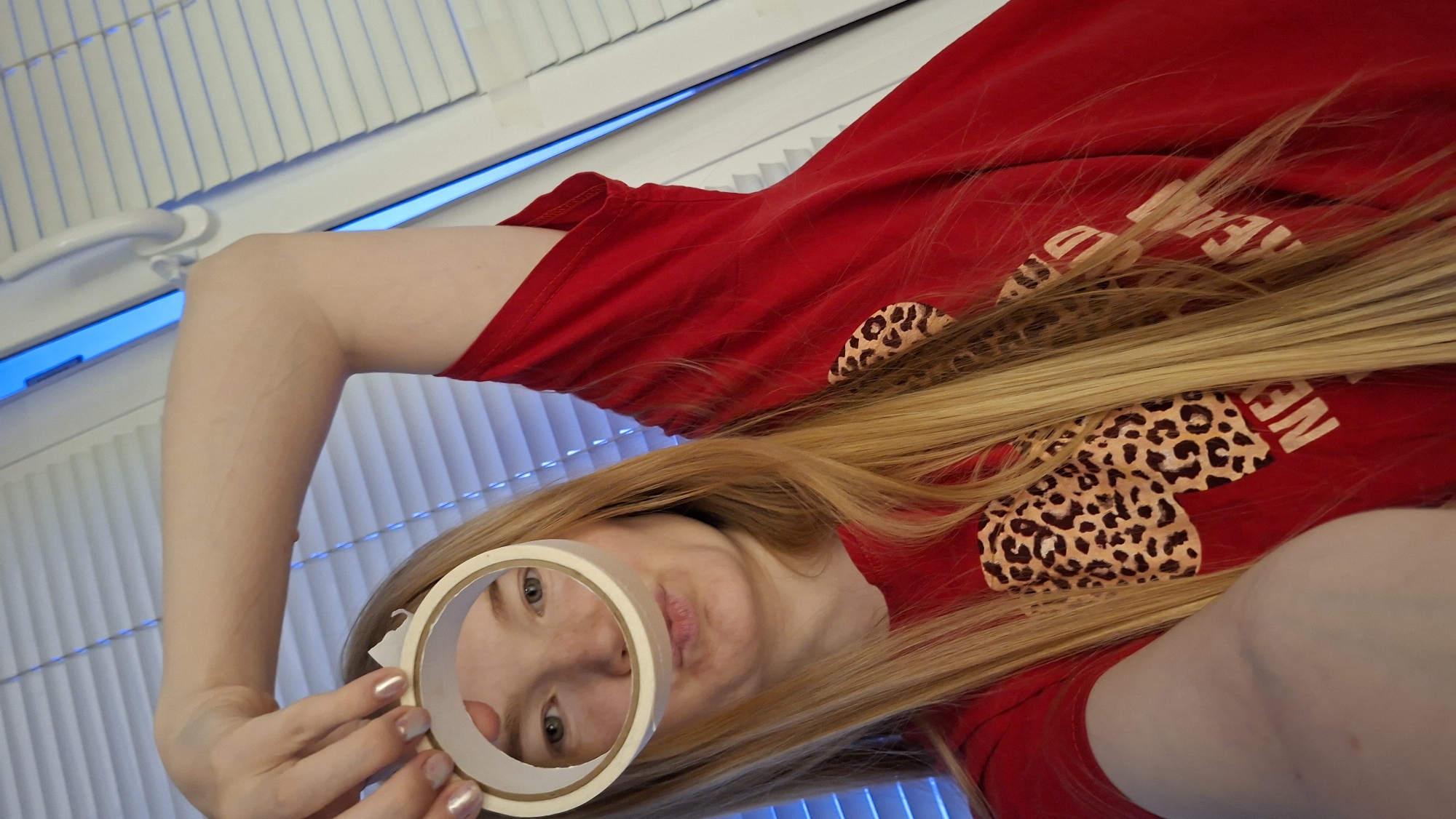 A person with long blonde hair and fair skin, wearing a red t-shirt with a leopard print design and white text, holds a roll of masking tape in front of their face. The background features white blinds and a white window frame with blue LED lighting. The location is in Chaikovsky, Russia.