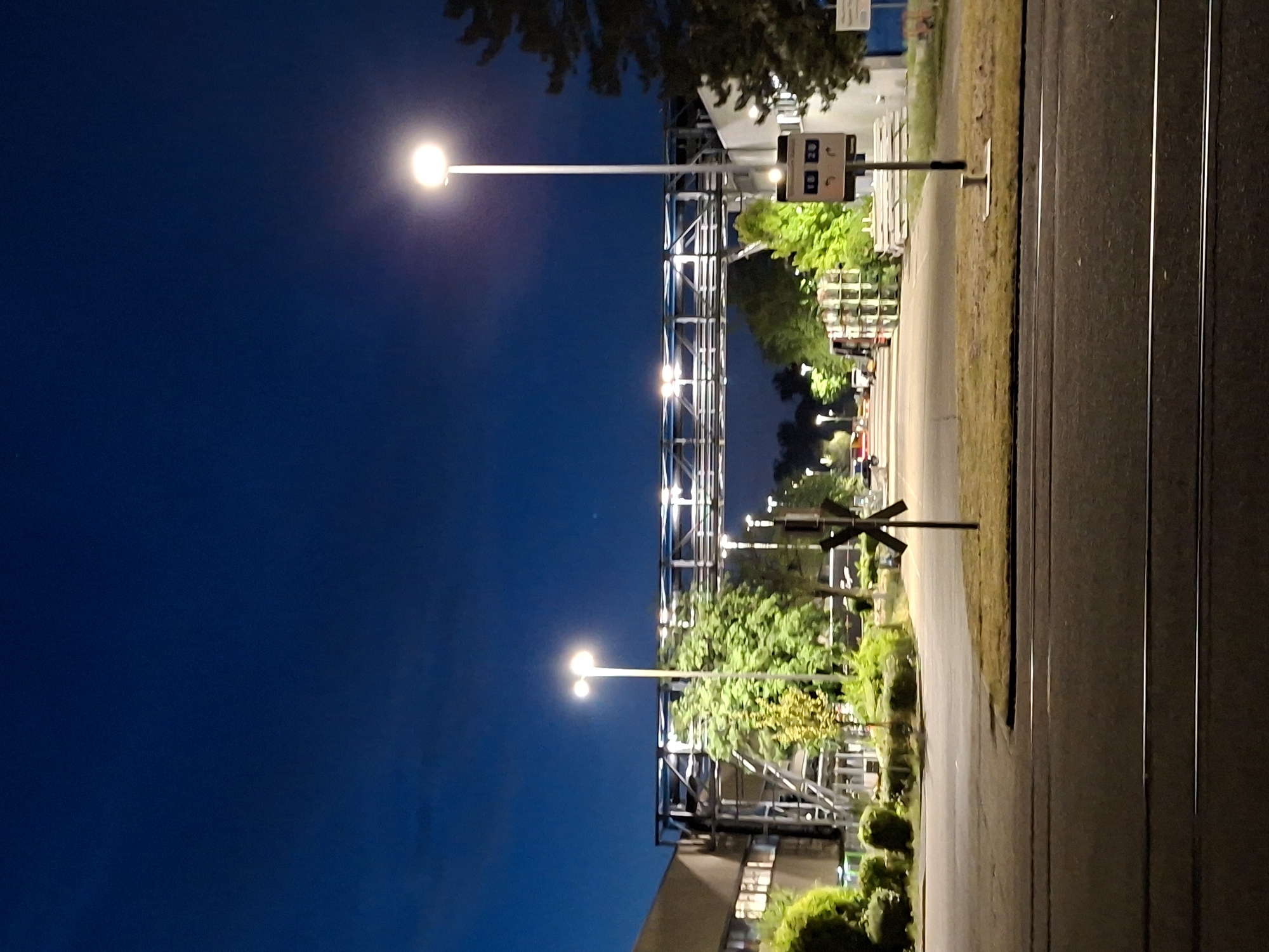 The image depicts an outdoor scene at dusk or night in the city of Rastatt, Germany. The sky is a deep, dark blue, indicating either twilight or a well-lit night. In the foreground, a paved road with what appear to be tram tracks runs horizontally across the frame.
Dominating the mid-ground is a long, elevated industrial walkway or bridge structure, supported by numerous metal poles and beams, spanning across a road or open area. This structure is illuminated by multiple bright lights. To the left of the walkway is a long, multi-story building with windows, suggesting an industrial or commercial facility.
Several streetlights are visible, casting pools of light. One prominent streetlight stands tall on the right side of the frame, near a small sign with the numbers "18 20" and two circular icons. To the left of the walkway, trees and lush green foliage line the area, some of which are also illuminated by artificial lights.
A crossbuck sign, typical of a railway or tram crossing, is positioned on the right side of the road in the mid-ground. Further down the road, more trees and lights are visible, and some vehicles, including what appears to be a truck, are present. The overall impression is one of an industrial or infrastructure area on the outskirts of a city, captured during a time of low natural light. There are no people visible in the image.